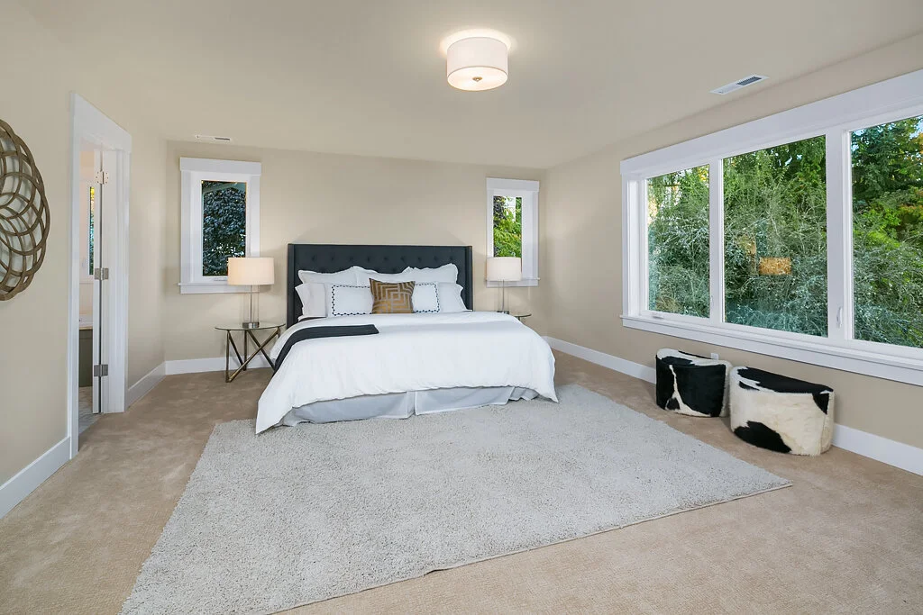 A bedroom with a large bed, black headboard, white bedding, and decorative pillows. There are two bedside tables with lamps, large windows showing greenery outside, and a small rug on tan carpet. Decorative storage boxes are on the floor near the win