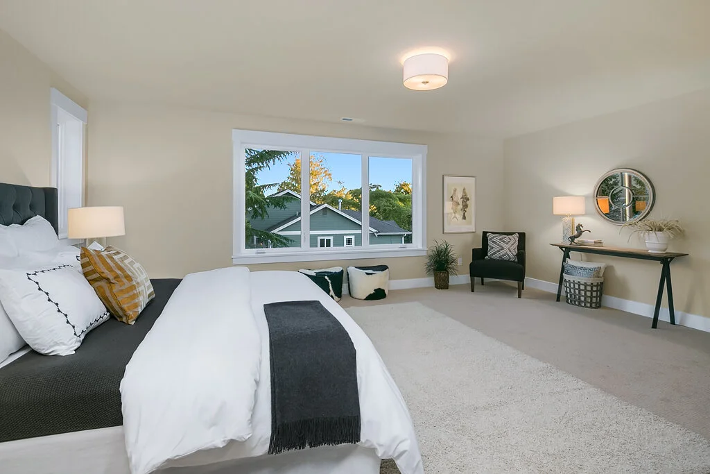A bedroom with a large window, a bed with white and patterned pillows, bedside lamps, a black chair, a round mirror, a console table, a plant, and a decorative basket, with neutral-colored walls and carpet.