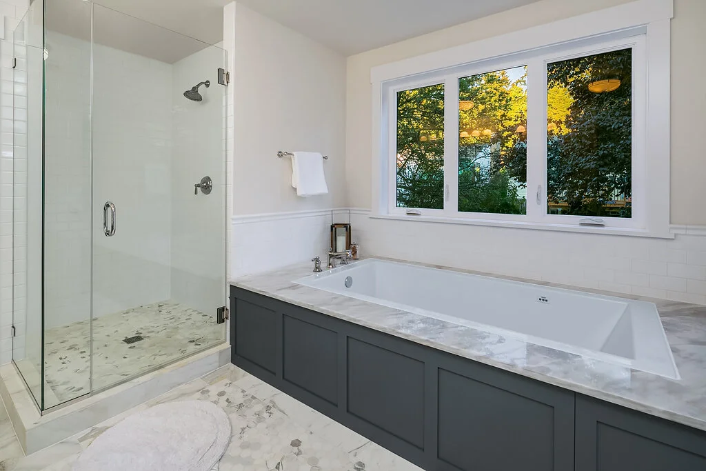 Bathroom with a walk-in glass shower and a large bathtub beneath a window showing a view of trees outside.