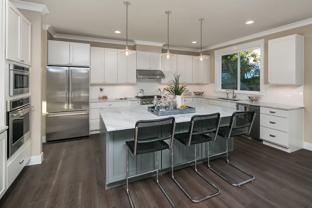 Modern kitchen with white cabinets, stainless steel appliances, a large kitchen island with a marble top, and three black bar stools. There is a window with a view of greenery.