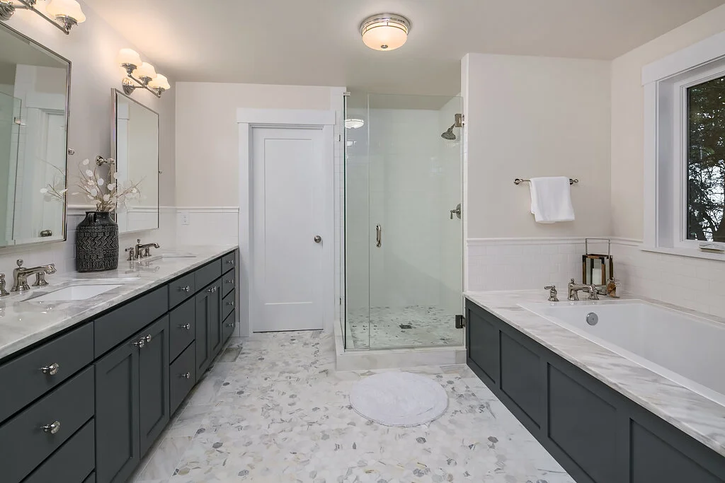 A clean, modern bathroom with double vanity sinks, dark gray cabinetry, a large bathtub, and a glass-enclosed shower with white tiled walls. There is a window with a view of trees and a white door.