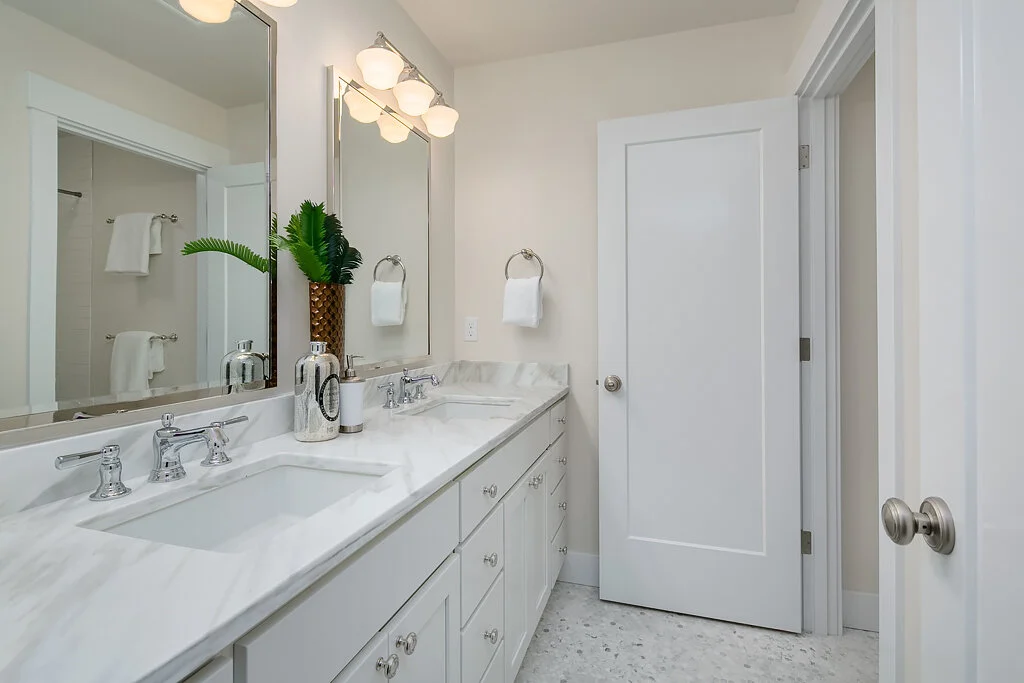 A bright bathroom with a double vanity with a marble countertop, two sinks, and chrome faucets. There are two large mirrors above the sinks, with a row of three lights above. Decor includes a potted plant, soap dispensers, and hand towels on both sid