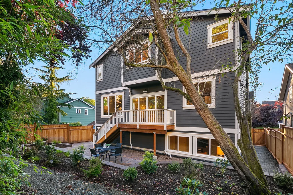 Backyard of a dark gray multi-story house with white trim, featuring a small wooden deck with white railing, a patio table with chairs, and a sloped garden area with plants and trees, bordered by a wooden fence.