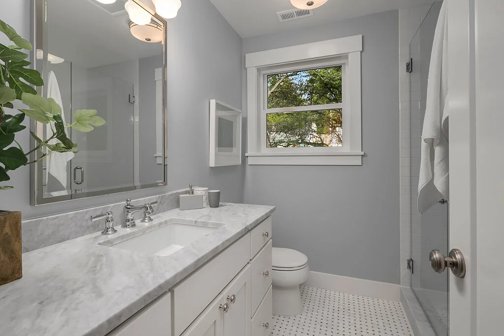 A modern bathroom with a marble countertop, a white sink, and silver fixtures. Features a large mirror, a window with a view of trees, a toilet, and a glass-enclosed shower. Decor includes a potted plant and bathroom accessories.