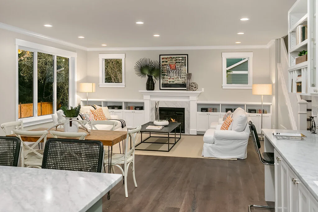 Living room with white walls, hardwood floors, a fireplace, built-in shelves, white sofas, a wooden dining table with white chairs, large windows, and modern decor.