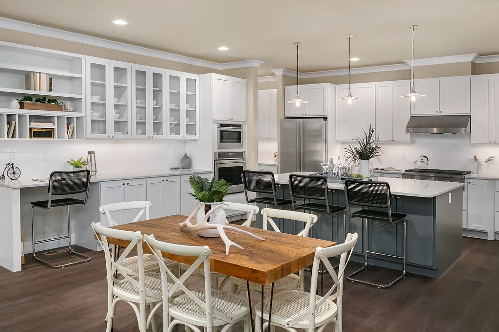 A modern, open-concept kitchen with white cabinets, stainless steel appliances, and a large island with seating for six. There is a wooden dining table with six white chairs in front of the kitchen. The space is decorated with plants and minimal acce