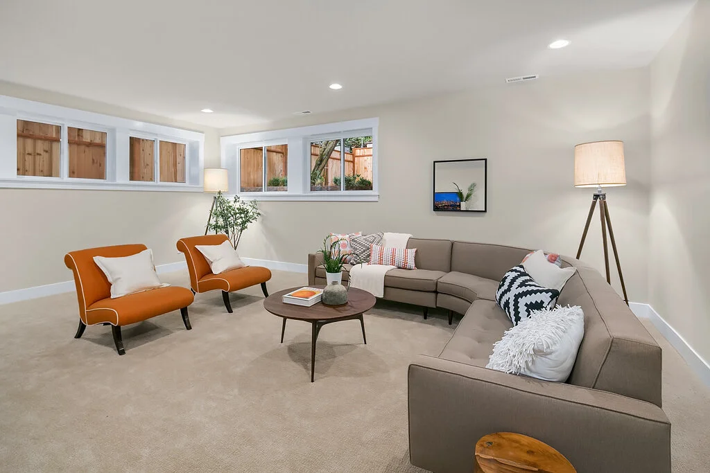 Living room with beige carpet, L-shaped sofa, two orange armchairs, wooden coffee table, floor lamps, and windows with a view of a wooden fence and greenery.