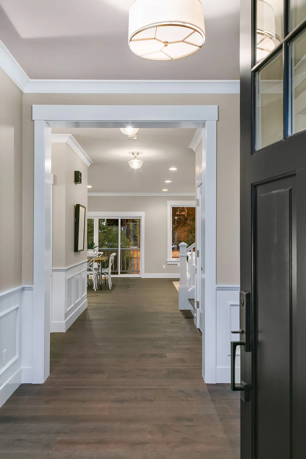 View from the front entrance into a modern dining area with large windows, hardwood floors, and white walls, featuring a black front door and ceiling lights.
