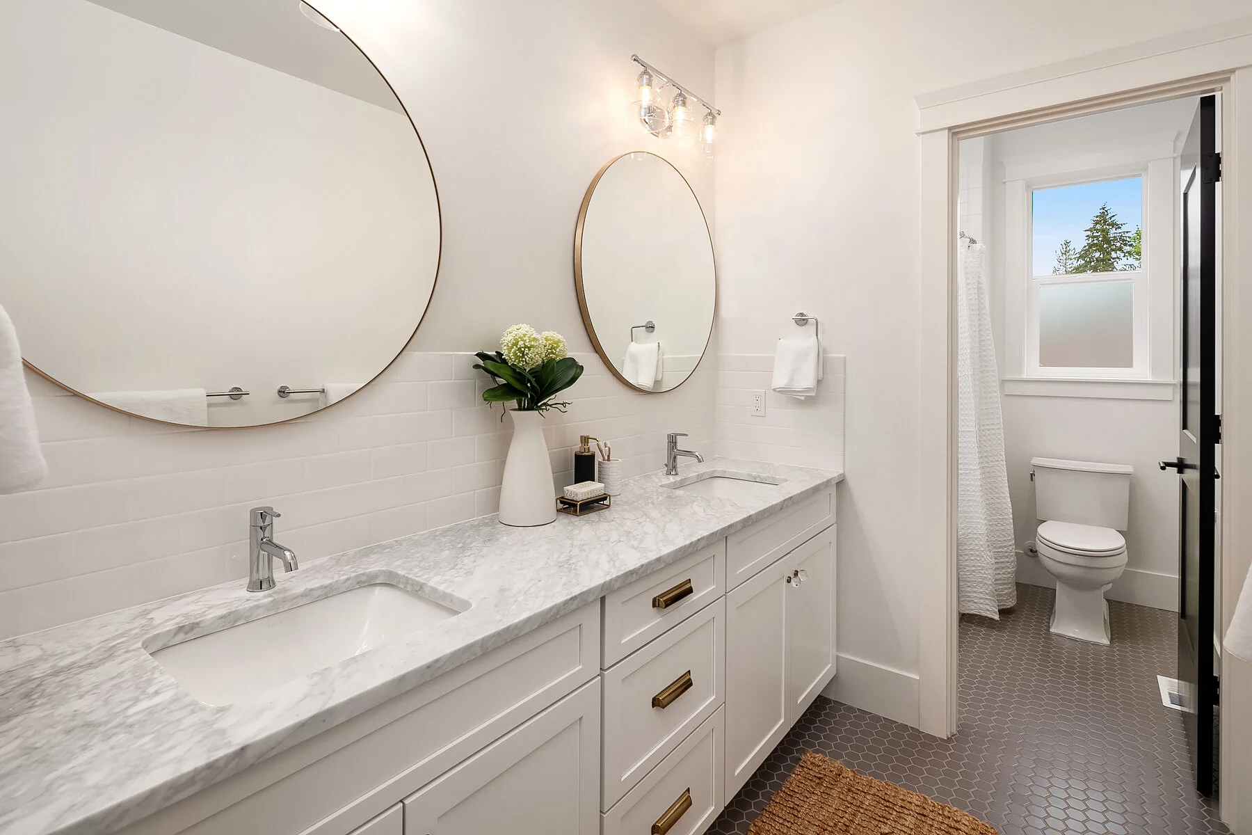 A modern bathroom with a double vanity featuring a marble countertop, two oval mirrors, and a vase with white flowers. In the background, there is a separate toilet area with a window and a white shower curtain.