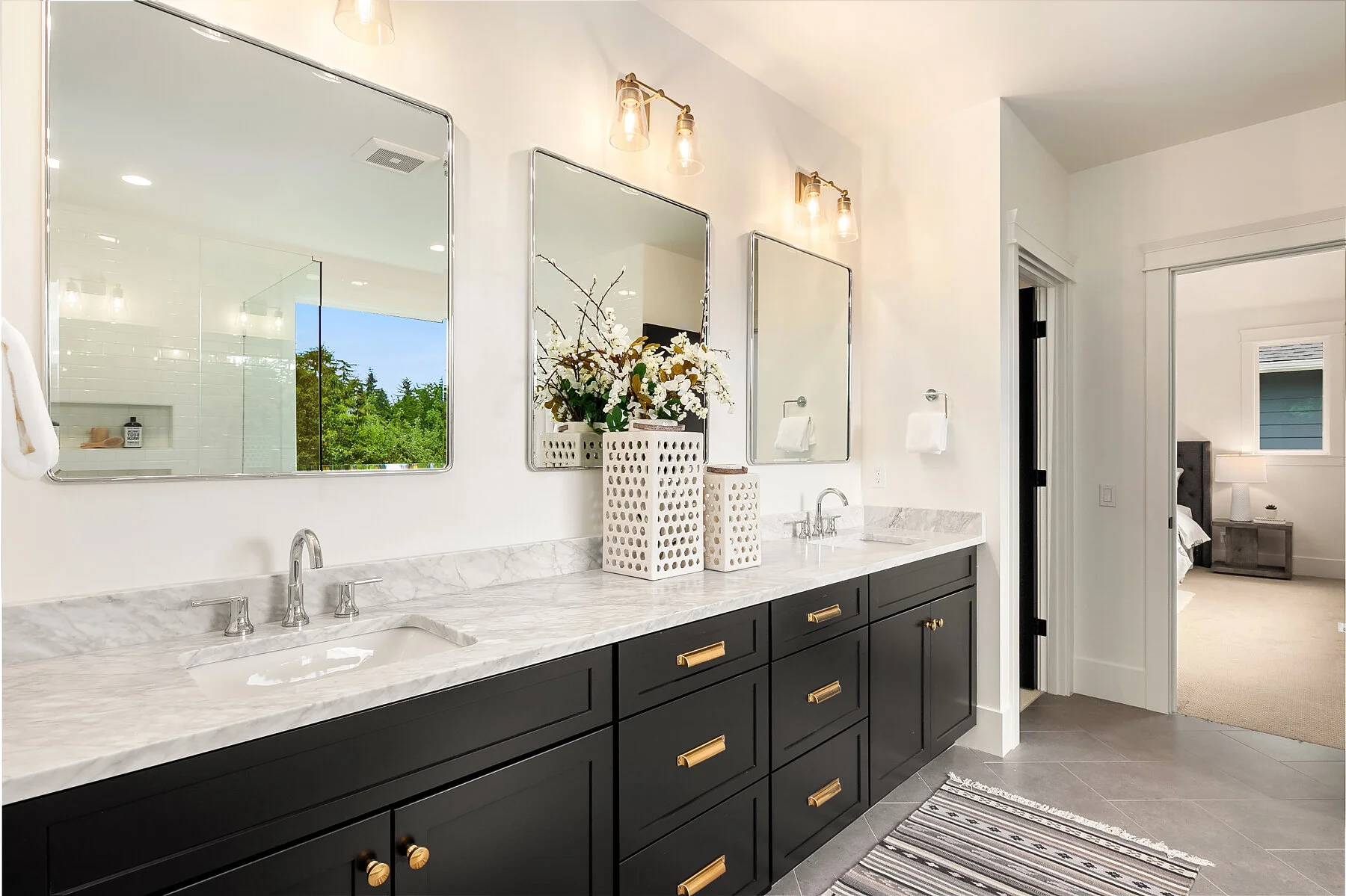 Bathroom with gray tiled flooring, black cabinetry with gold handles, marble countertops, three rectangular mirrors, and three wall-mounted lights. A door opens to a bedroom with a nightstand and bed visible.