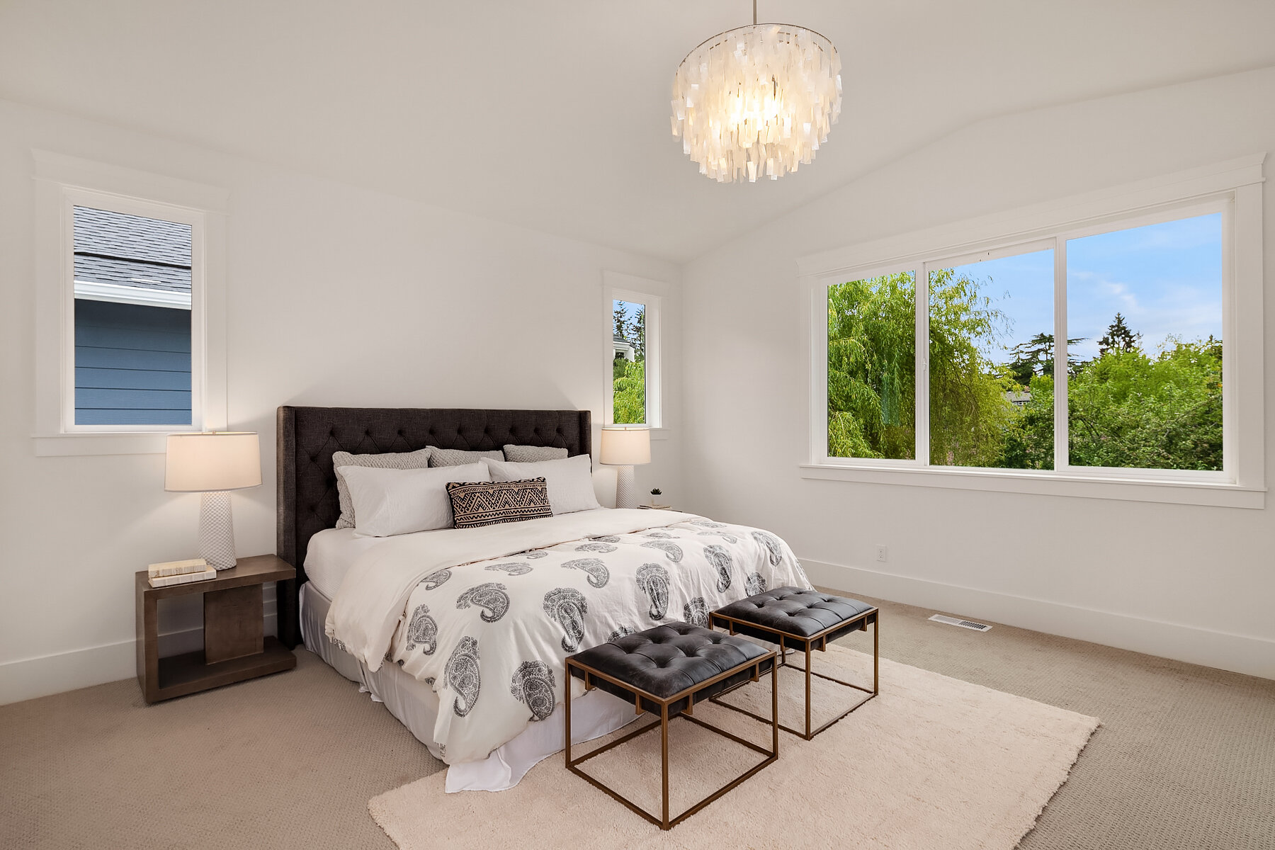 Bright bedroom with large windows showing green trees outside. The room has a dark upholstered bed with white bedding and a decorative pillow. Two matching bedside tables with lamps, and two black tufted benches at the foot of the bed. A modern chand