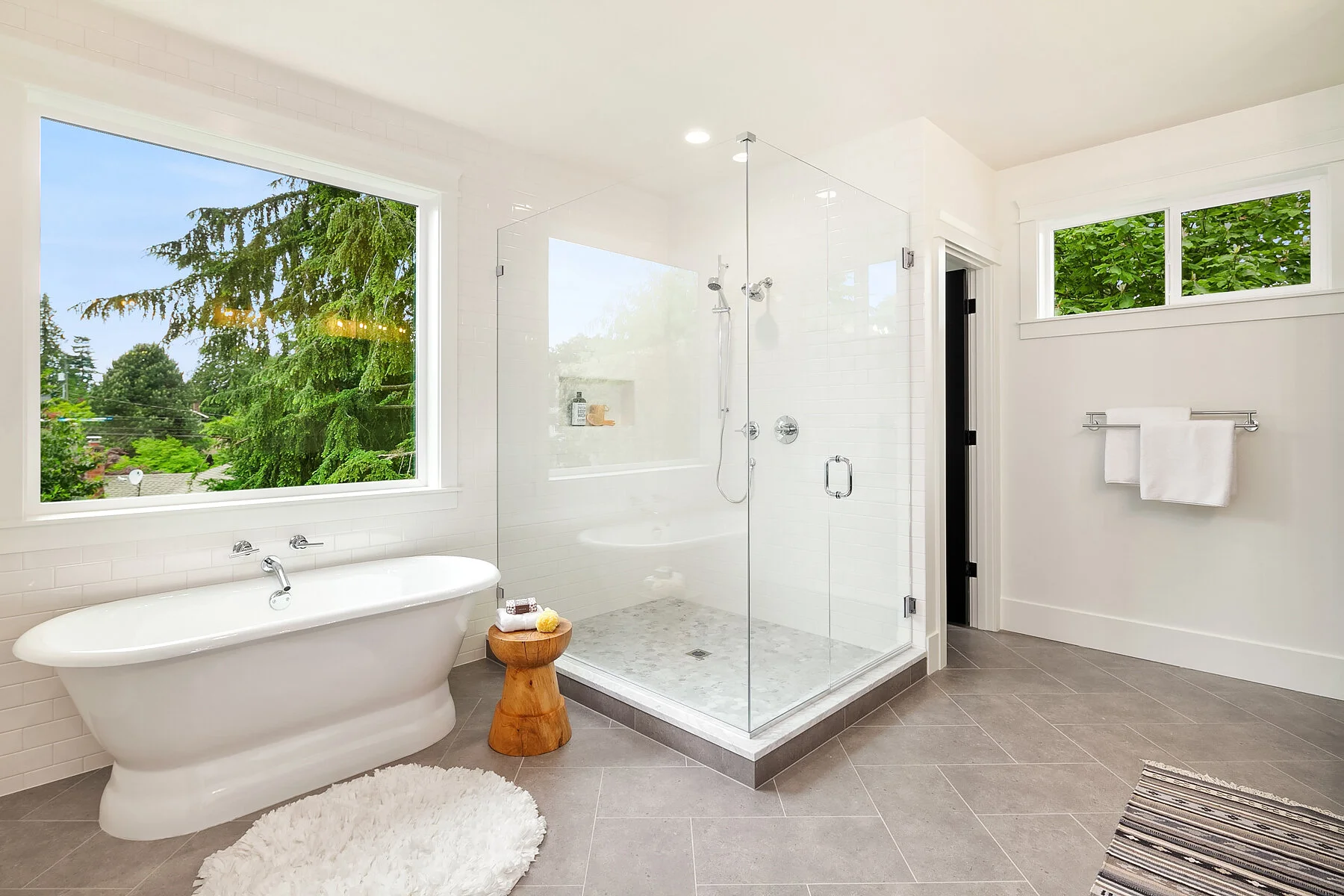 Bright modern bathroom with large windows, a bathtub, a glass-enclosed shower, and gray tiled flooring.