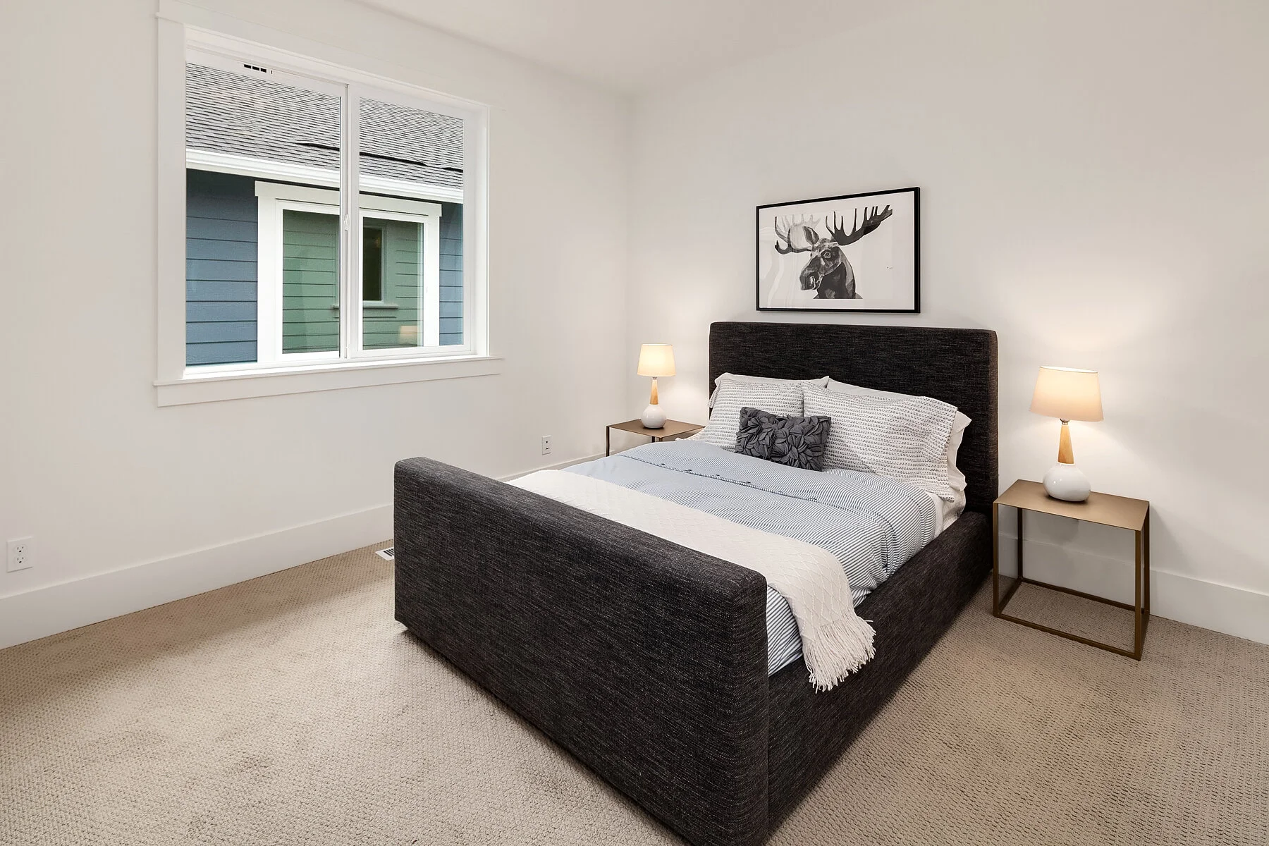 A bedroom with a dark upholstered bed, white and gray bedding, and a black and white moose artwork above the headboard. There are two small side tables with lamps on either side of the bed. A window shows a neighboring house with blue siding.