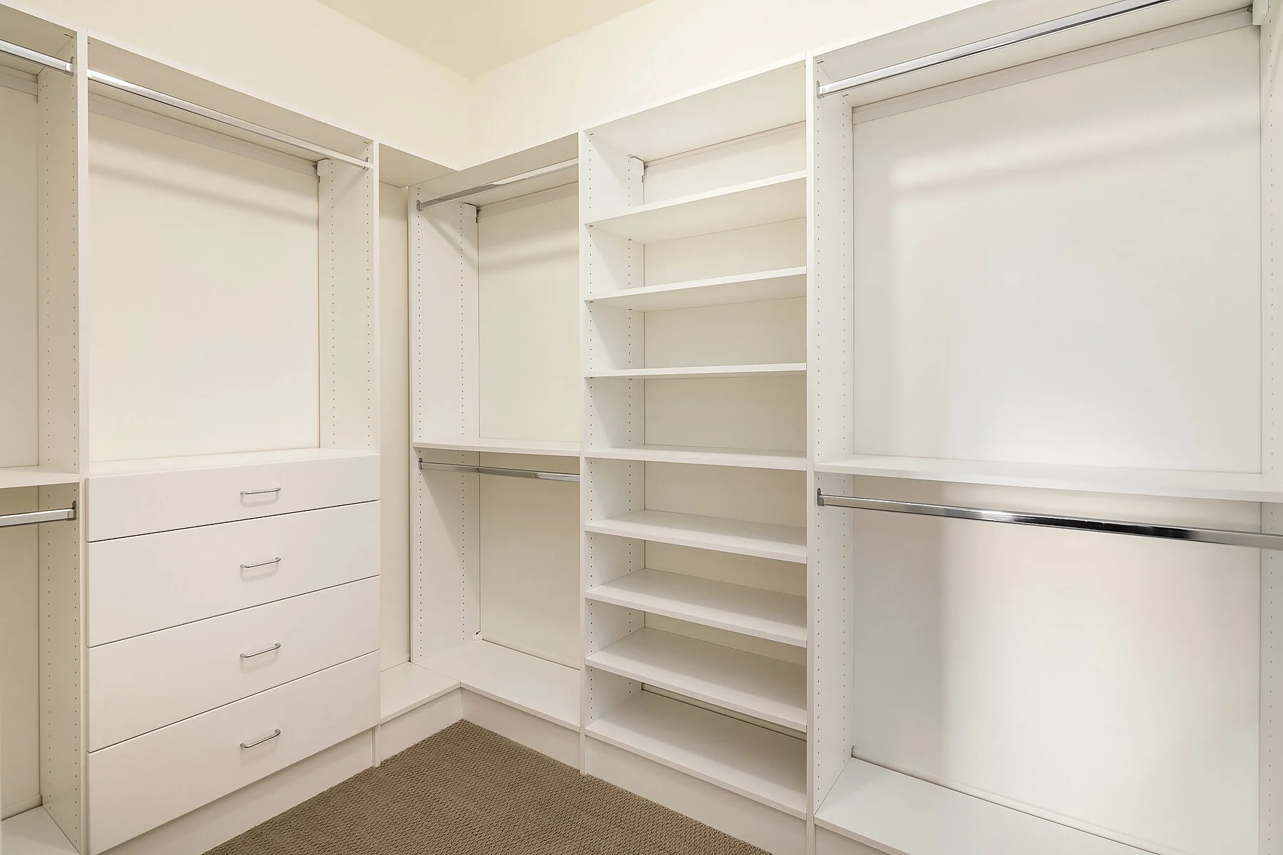 Empty white closet storage system with shelves, drawers, and hanging rods, on beige carpeted floor.