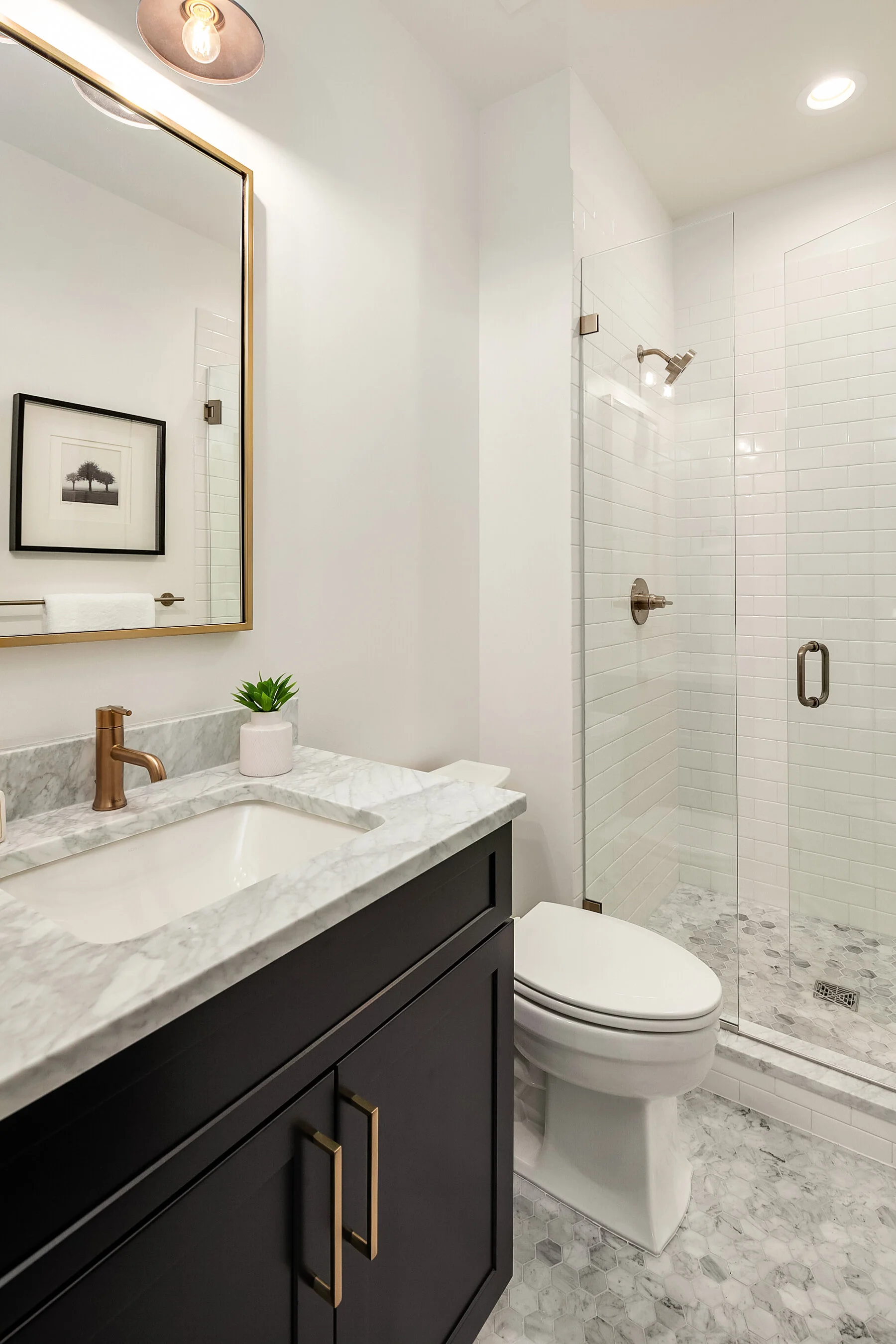Modern bathroom featuring a marble countertop vanity with a small green plant, a framed black-and-white artwork on the wall, a white toilet, and a walk-in shower with white subway tiles and a glass door.