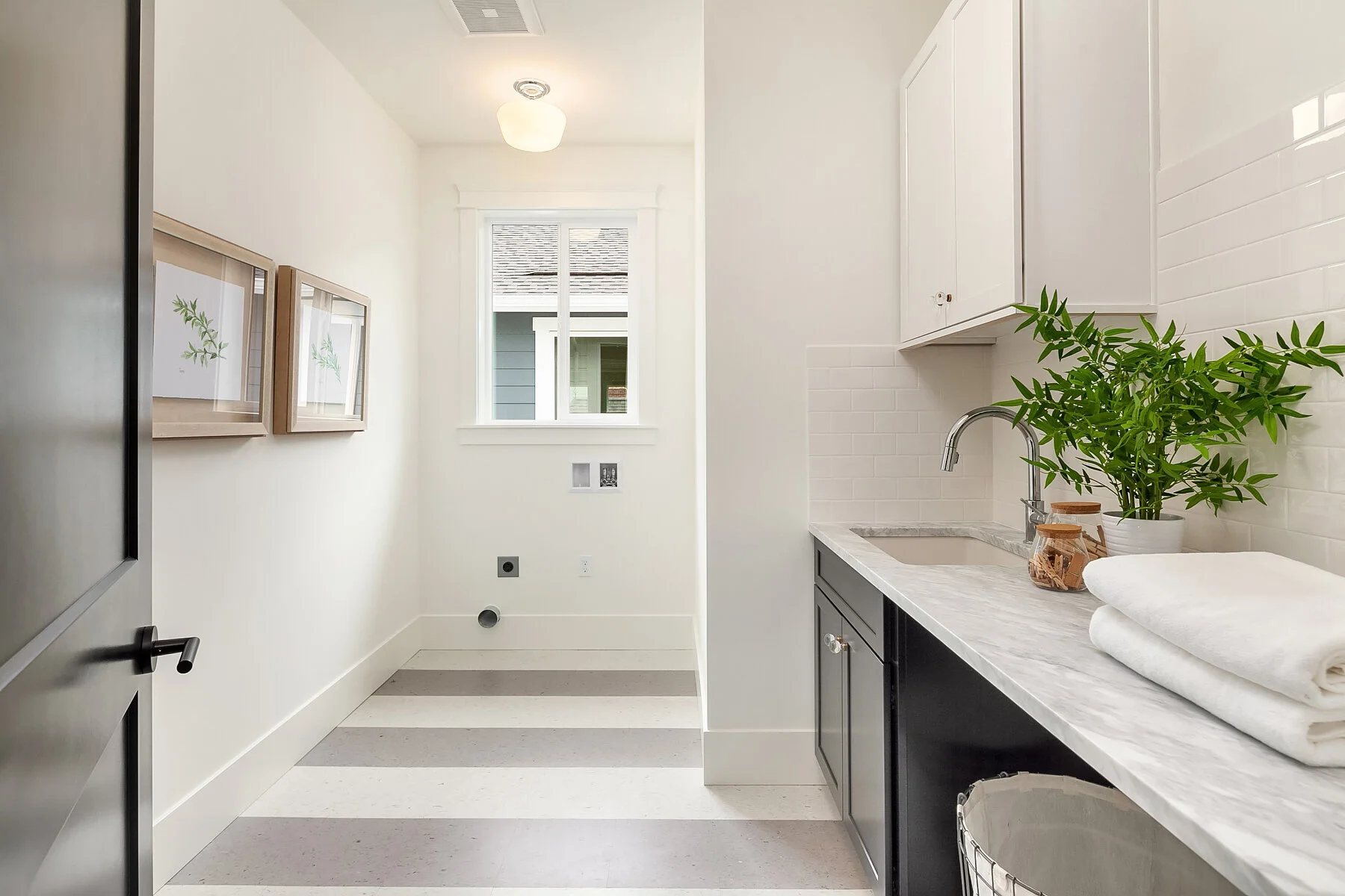 A bright laundry room with a white wall, a window, black and white cabinet, a potted green plant, and neatly folded white towels on a marble countertop.