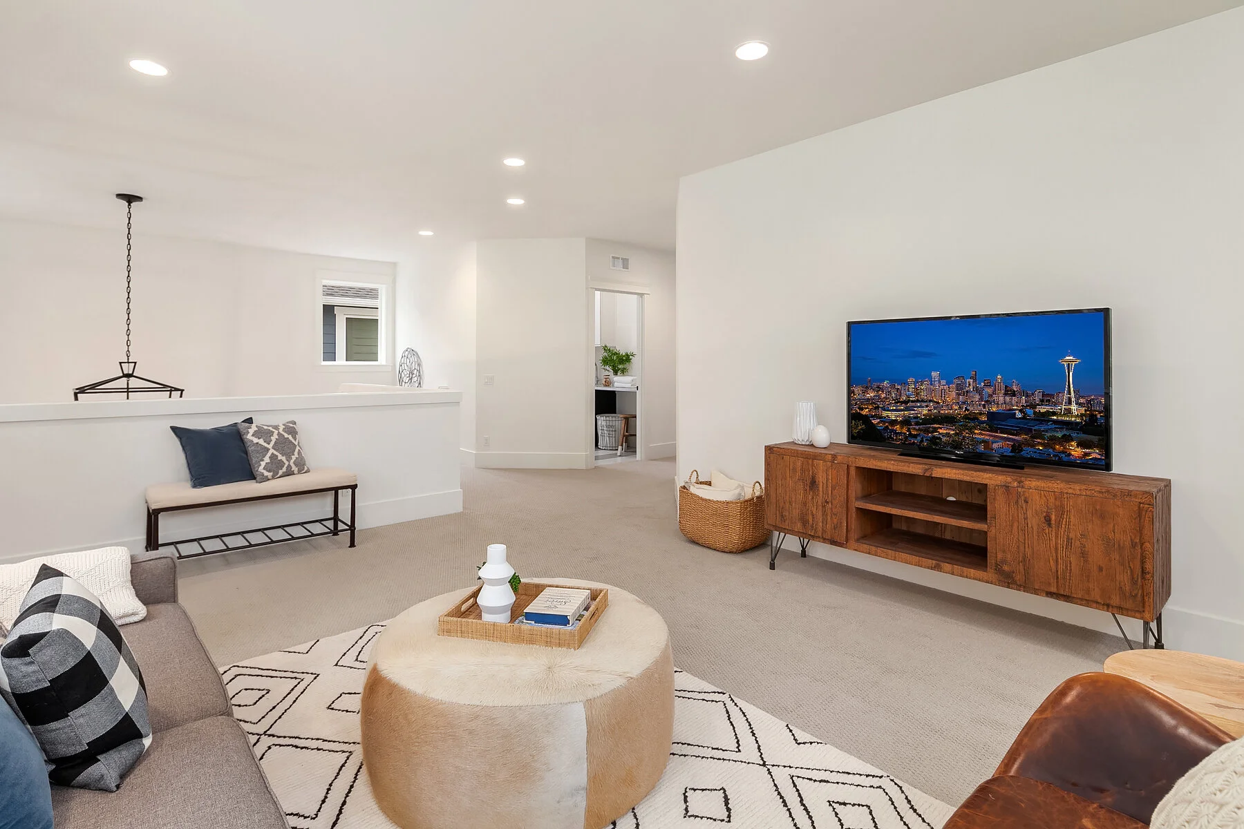 Modern living room with neutral tones, a beige sofa with plaid pillows, a round beige ottoman, a wall-mounted TV on a wooden media console, and a woven basket, with a view of downtown Seattle and the Space Needle visible on the TV screen.