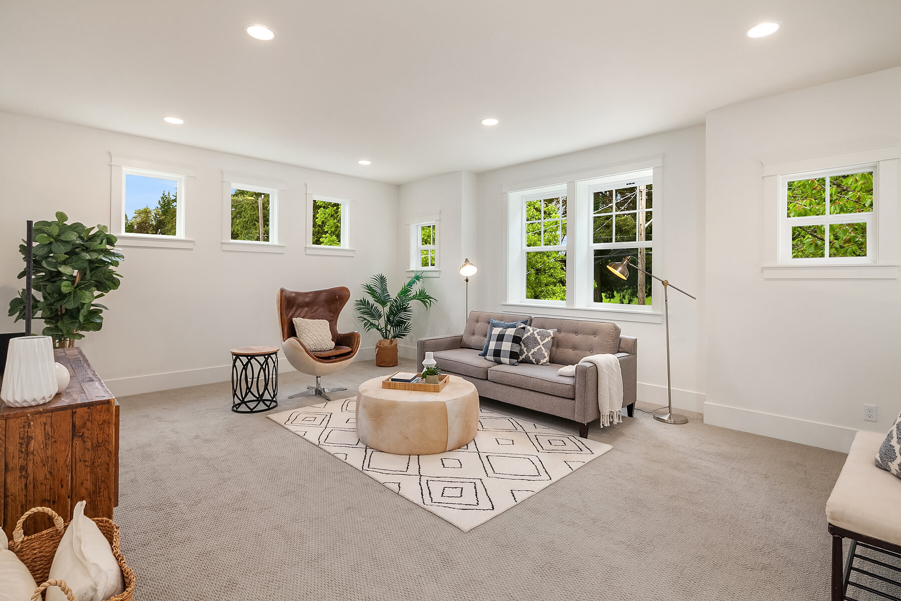 Bright living room with white walls, large multi-pane windows showing green trees outside, beige carpet, a gray sofa with pillows, a modern standing lamp, a beige ottoman with a tray, a brown leather chair, a potted plant, and a wooden console with d