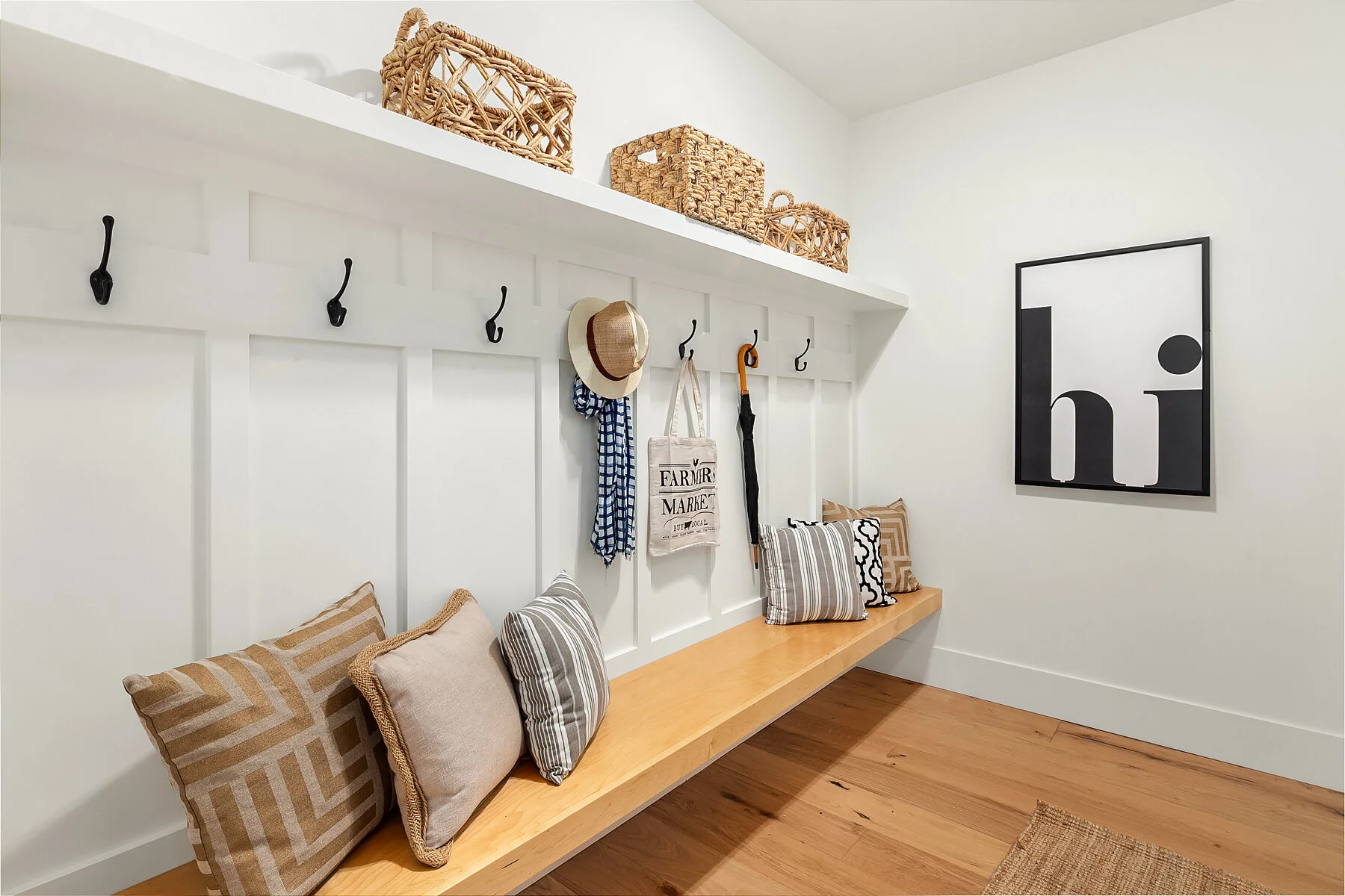 A minimalistic entryway with a white wall featuring black hooks, a wooden bench with striped and solid pillows, a framed art piece with the word 'hi' in black and white, and a wooden shelf with woven baskets above.