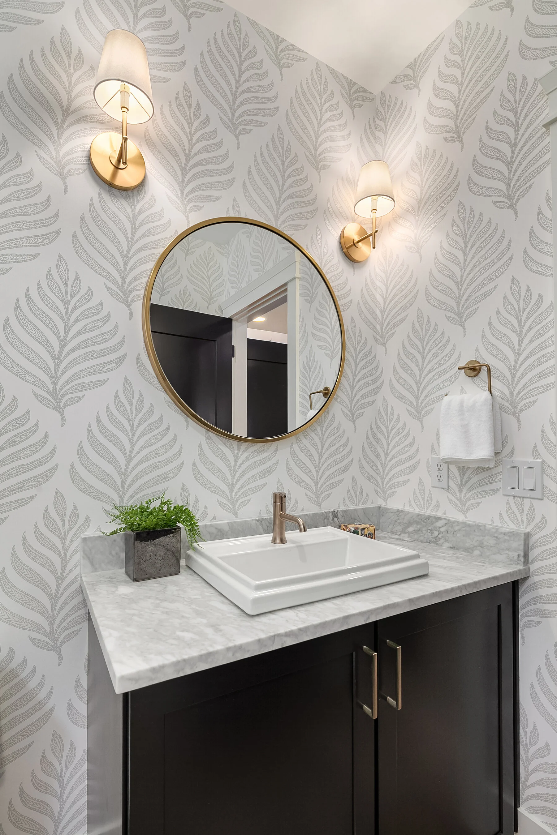 Bathroom vanity with a rectangular vessel sink, a marble countertop, black cabinet, circular mirror, wall-mounted light fixtures, towel, and small potted plant