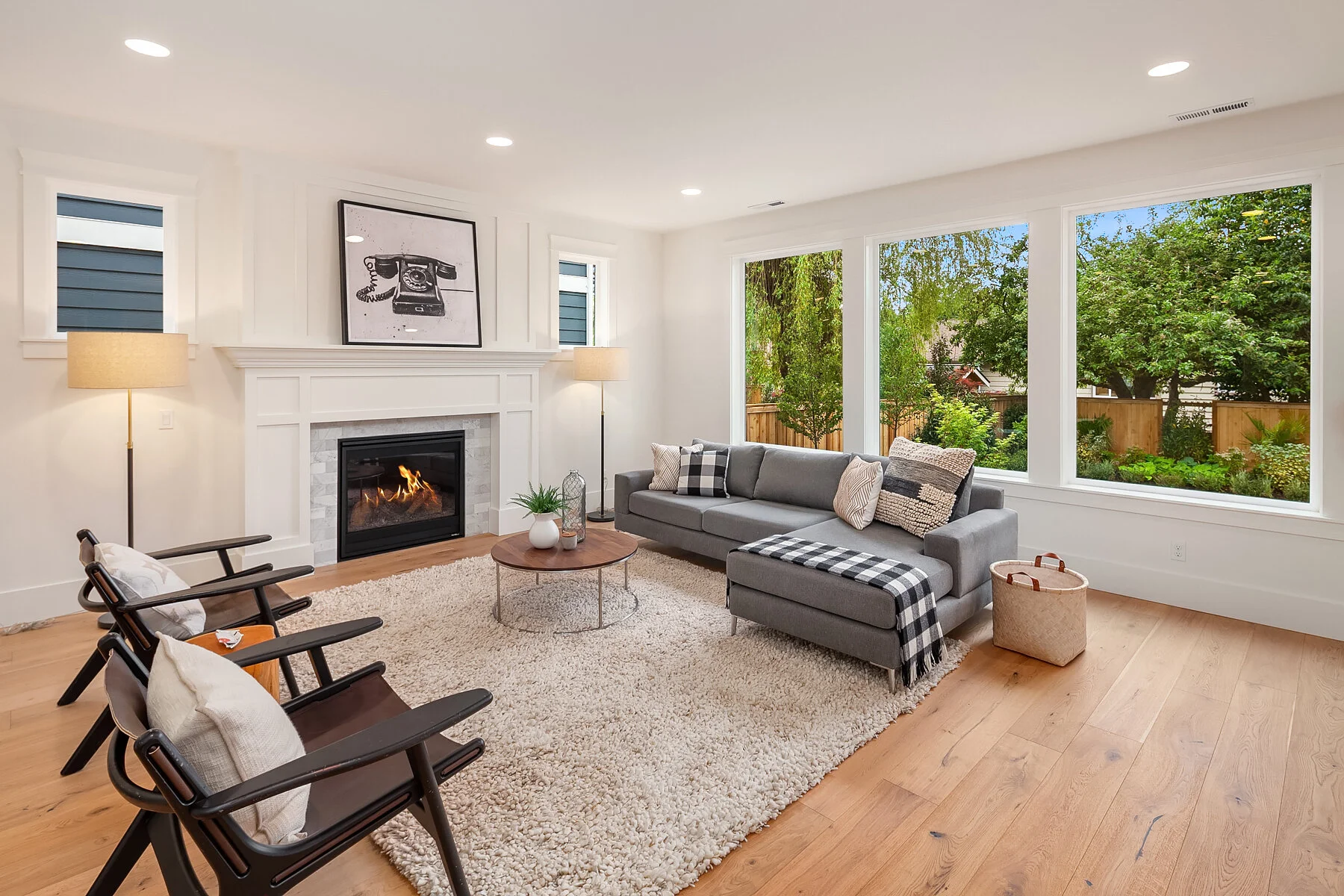 Bright living room with large windows, a gray sectional sofa with patterned pillows, a fireplace with a framed picture above, two black chairs with pillows, a round wooden coffee table, a beige rug, and outdoor greenery visible through the windows.