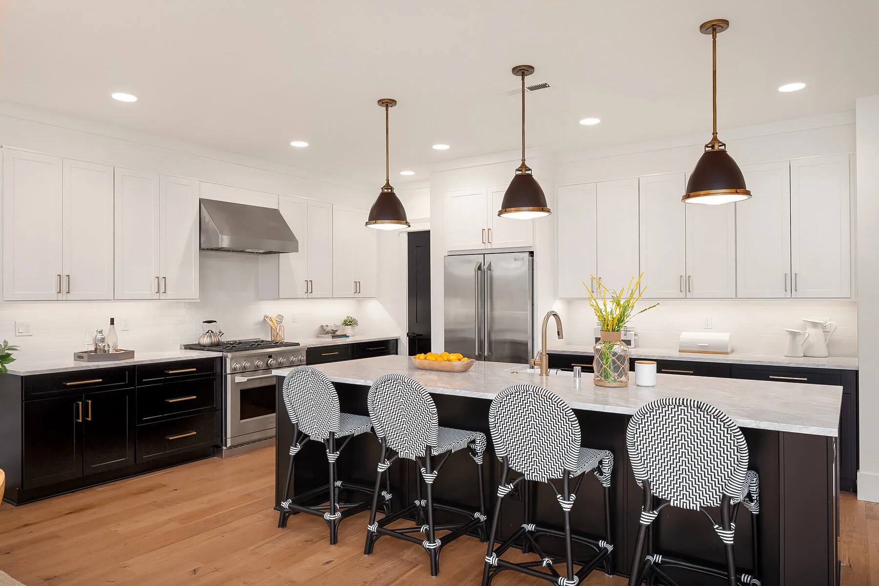 Modern kitchen with white upper cabinets, black lower cabinets, a marble countertop island, and three black and white patterned chairs. Stainless steel appliances include a refrigerator and oven, with gold fixtures and pendant lights over the island.