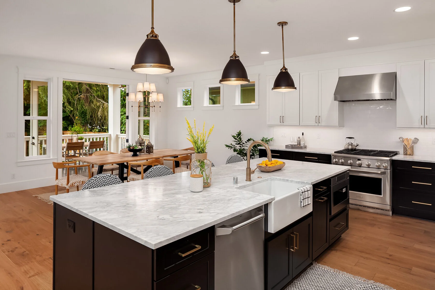 Modern kitchen with black cabinets, white marble island, stainless steel appliances, black and gold pendant lights, and a dining area with a view to a balcony outside.
