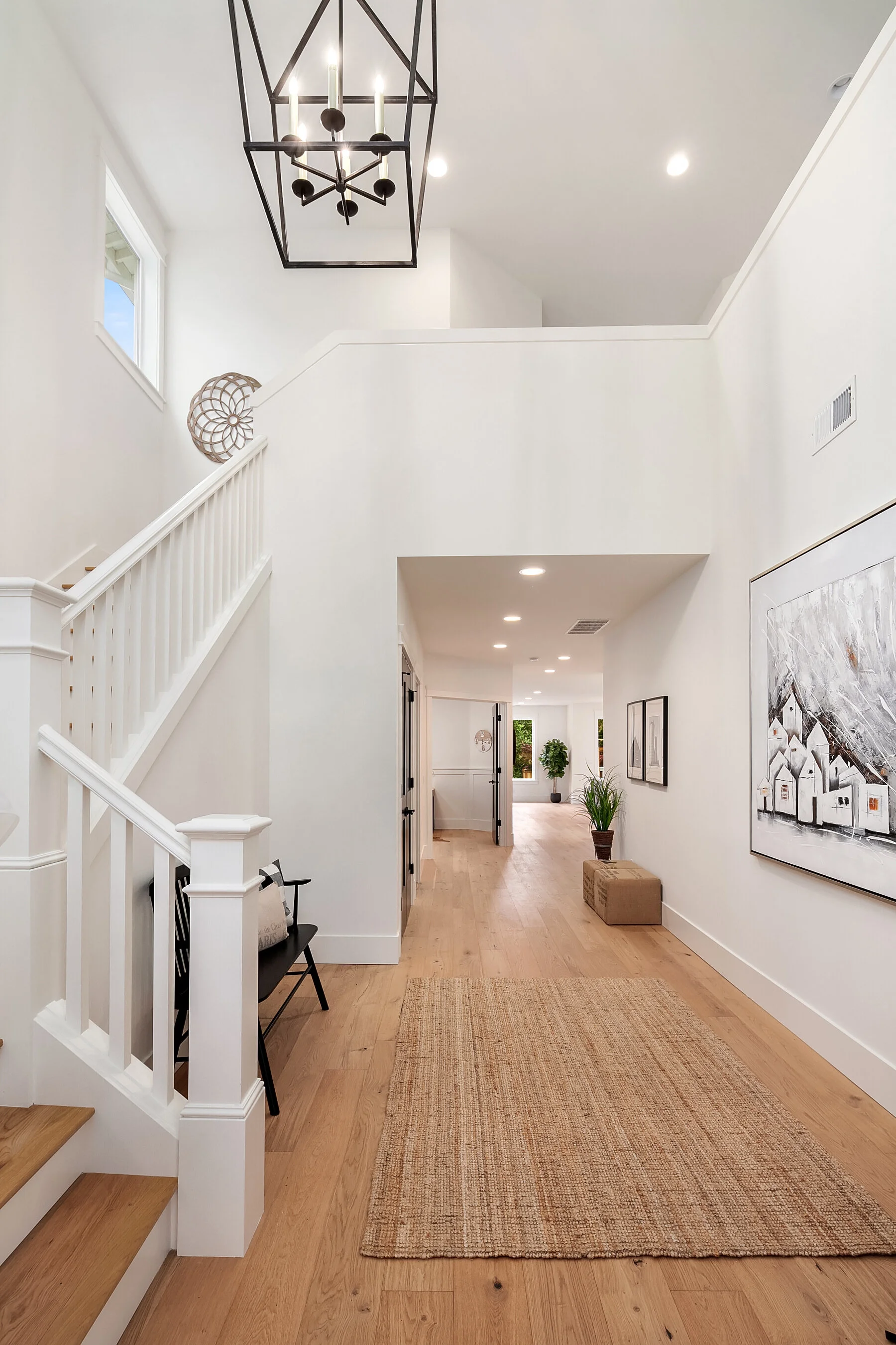 Bright, modern foyer with white walls, hardwood floors, and a beige rug. Features stairs to the left with black railing and hanging light fixture. Artwork on the right wall, potted plants towards the back, and open doorway leading to other rooms.