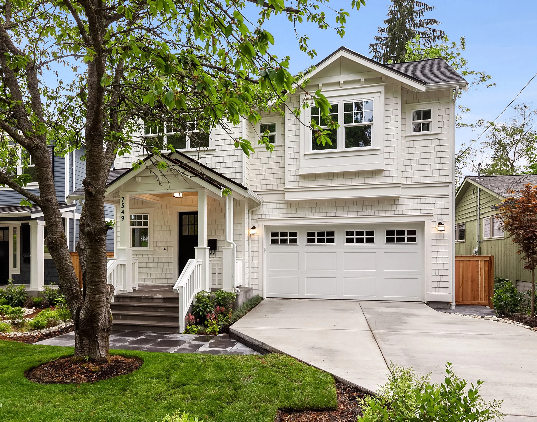 A two-story white house with a front porch, a black door, a two-car garage, and landscaped front yard with green grass, a large tree, and shrubbery.