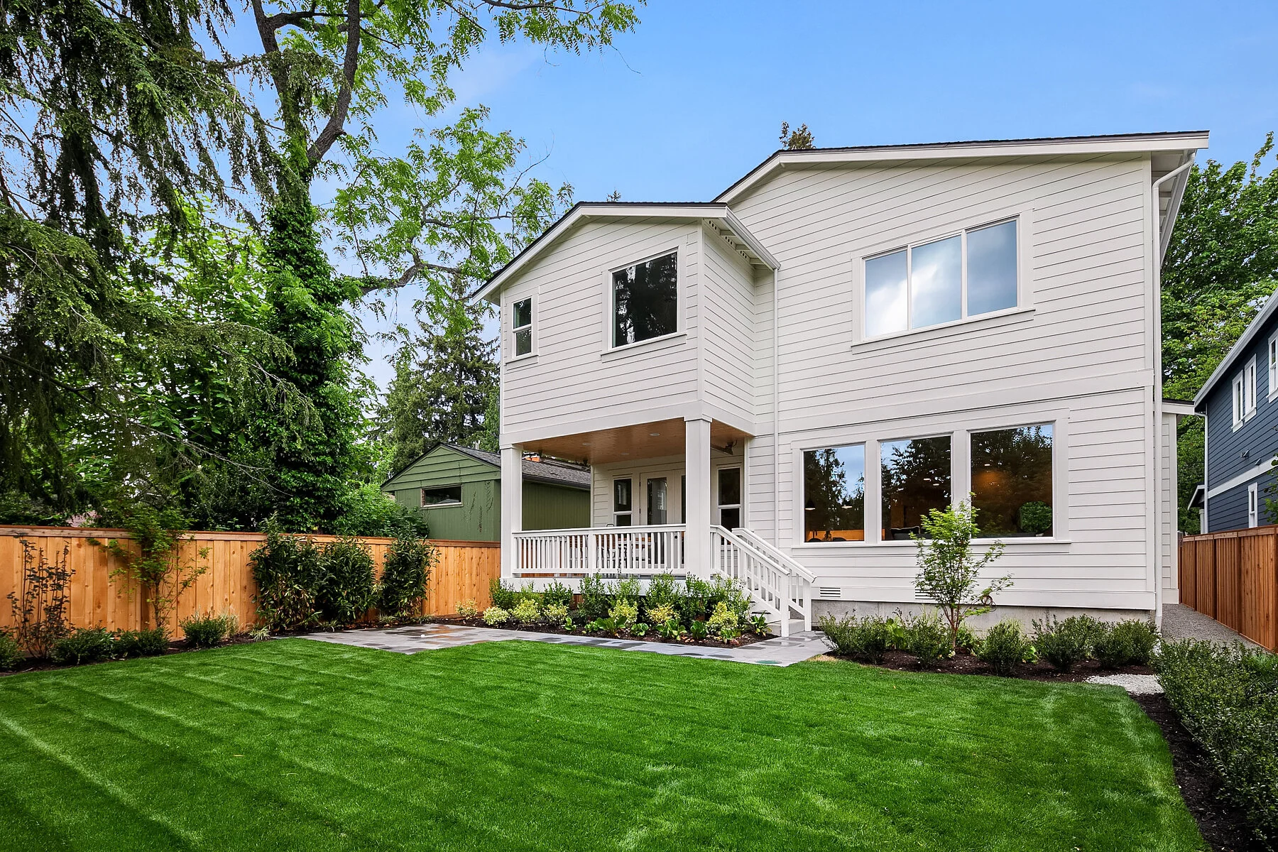 Front view of a modern white two-story house with a small covered porch, surrounded by well-manicured green lawn and garden, with tall trees and a blue sky in the background.