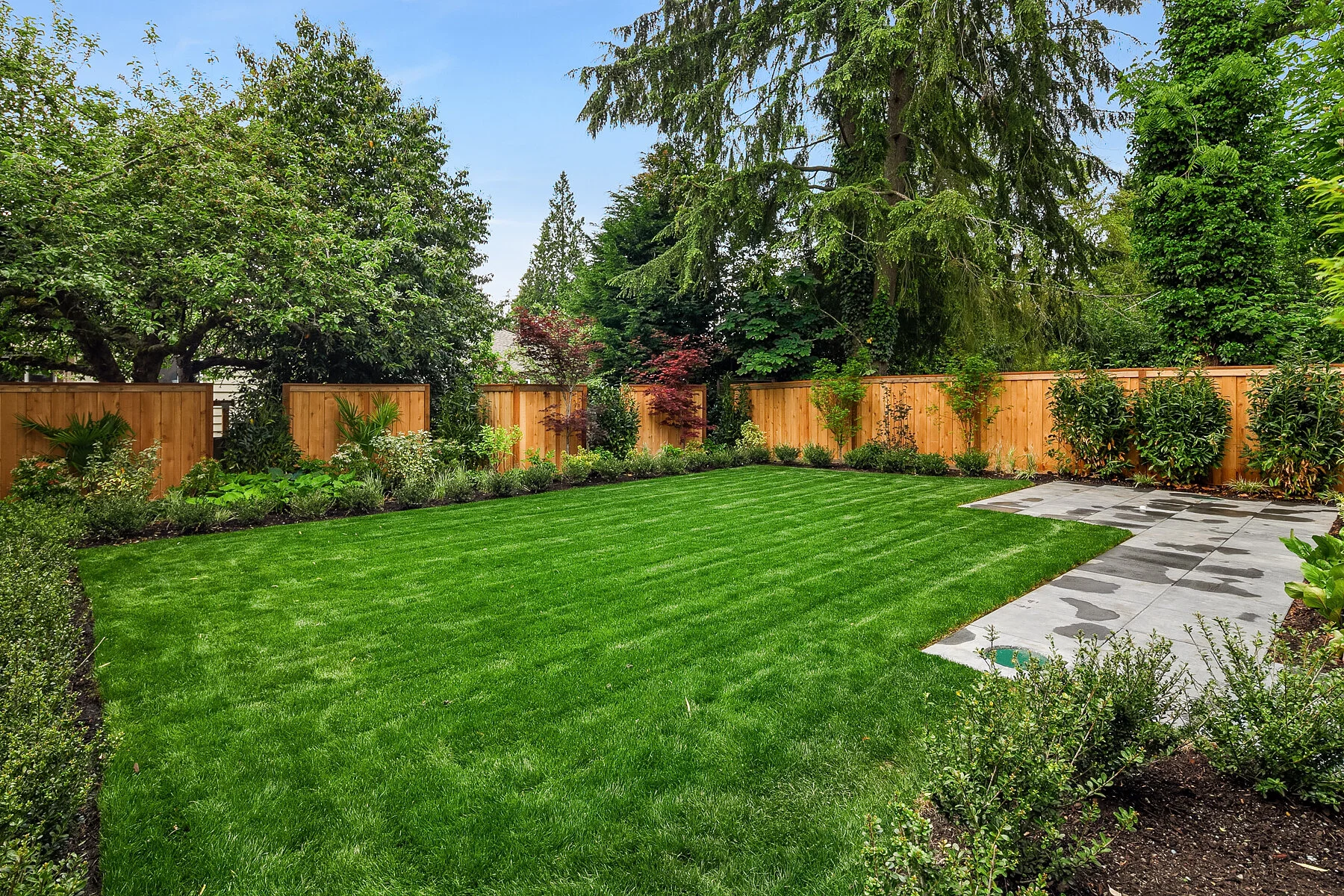 Well-maintained backyard with lush green lawn, flower beds along the fence, and a stone patio in the corner, surrounded by tall trees and a wooden privacy fence.