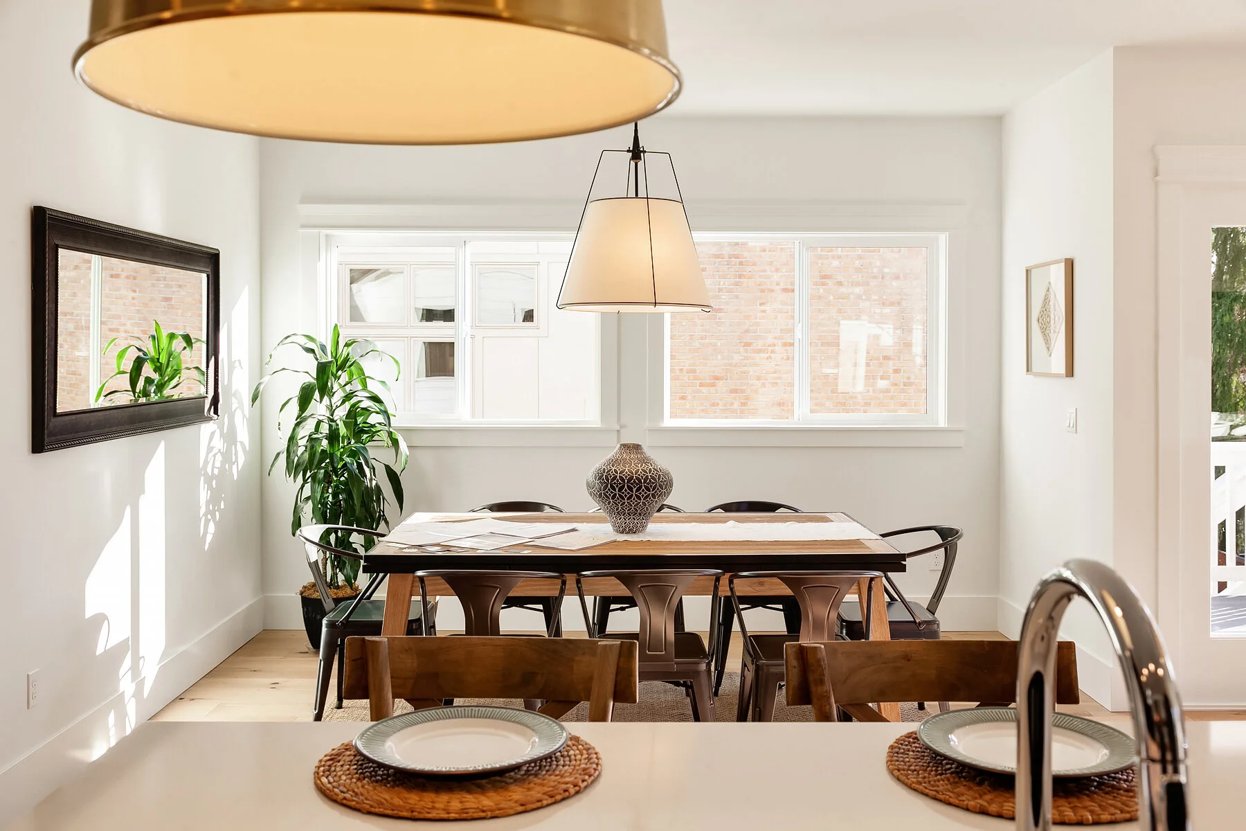 A dining room with a wooden table, six chairs, a decorative vase, and two pendant lights hanging from the ceiling. The room has large windows, white walls, and a potted plant in the corner.