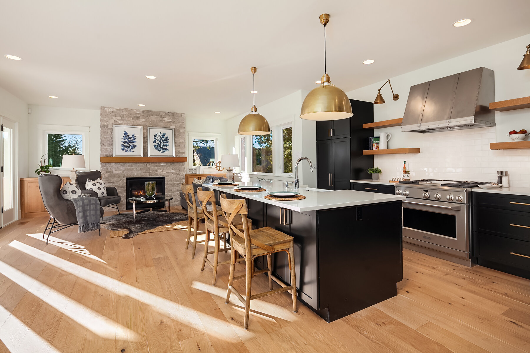 Open-concept kitchen and living room with black cabinetry, stainless steel appliances, a white island with wooden barstools, and a cozy seating area with chairs, a fireplace, and artwork on the wall.