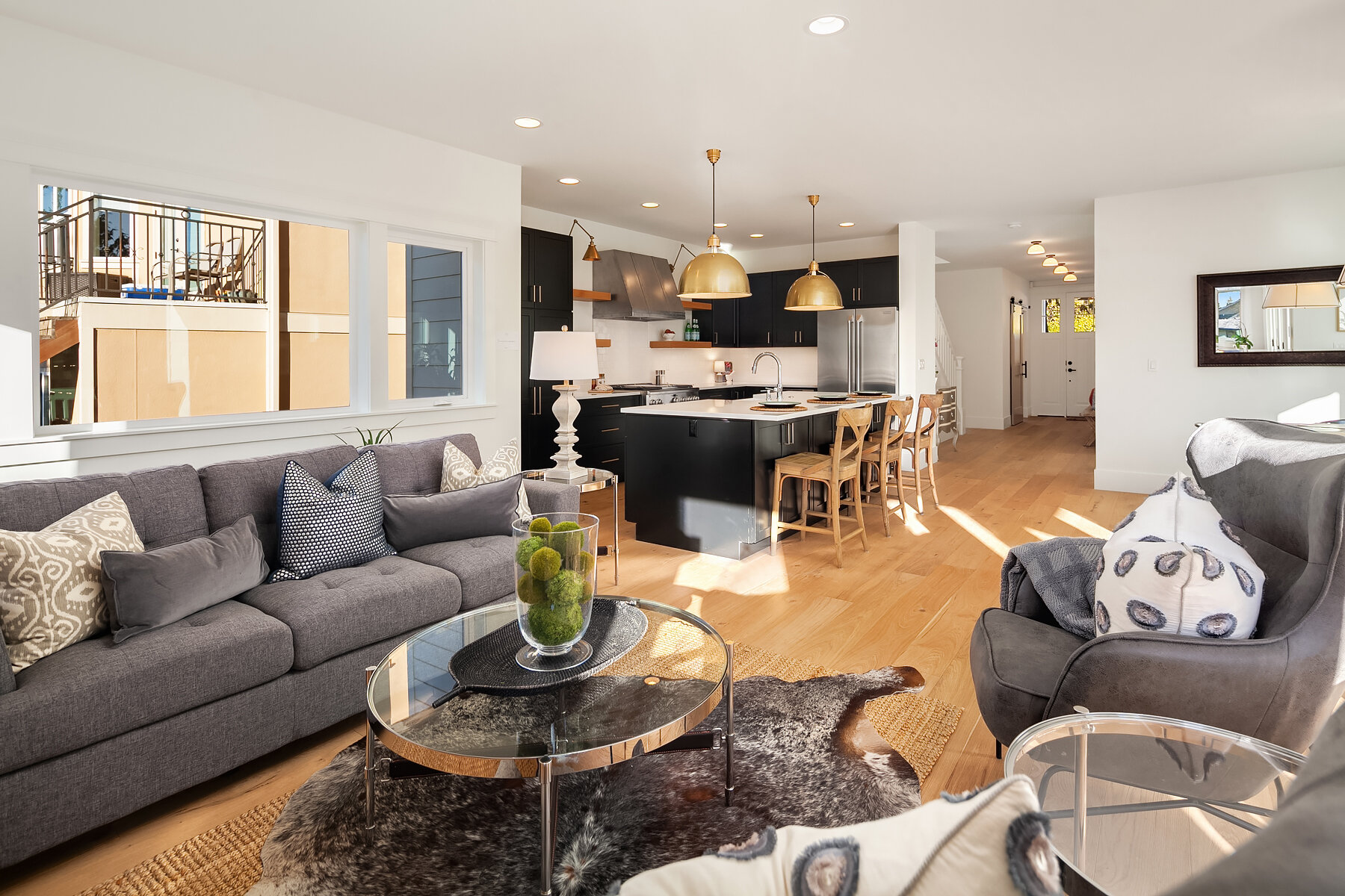 Open-concept living room and kitchen with gray sofa, armchair, glass coffee table with decorative greenery, black cabinets, kitchen island, wooden barstools, and sunlight streaming in.