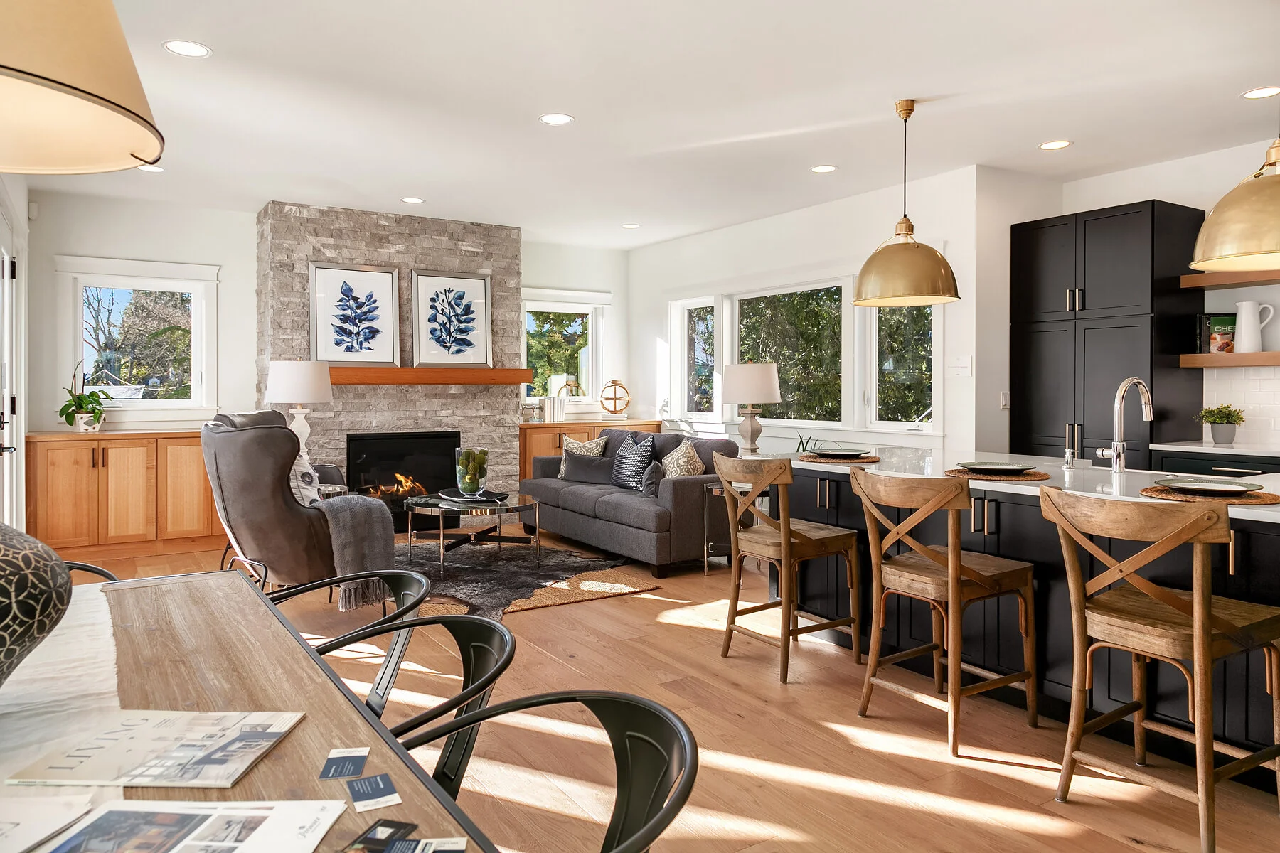 Open-concept living room and kitchen with natural light, modern decor, gray sofa, black cabinets, wooden barstools, and fireplace.