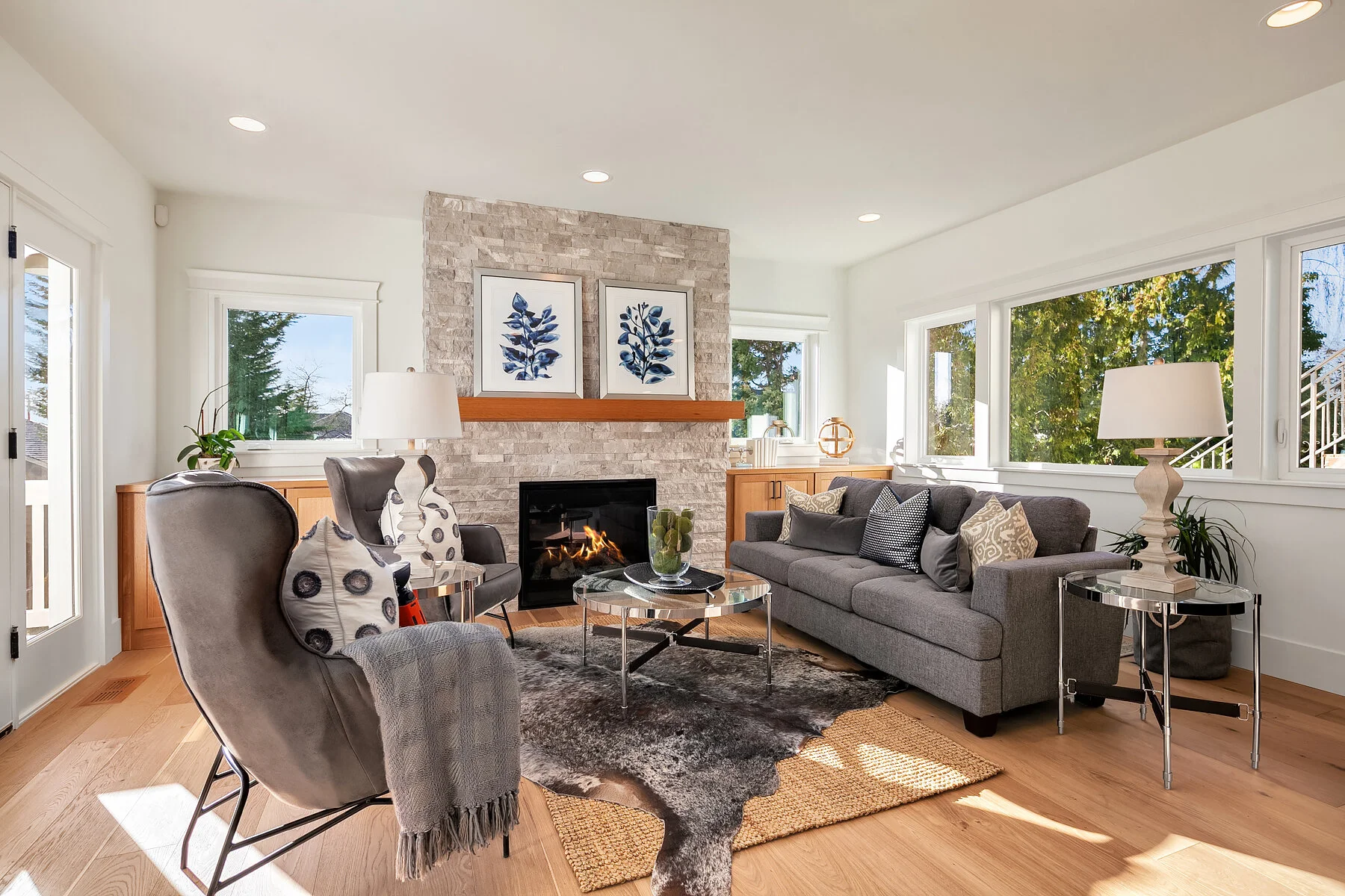 Living room with gray sofa, armchairs, and a fireplace, illuminated by large windows with trees outside, decorated with framed blue botanical prints and modern lamps.
