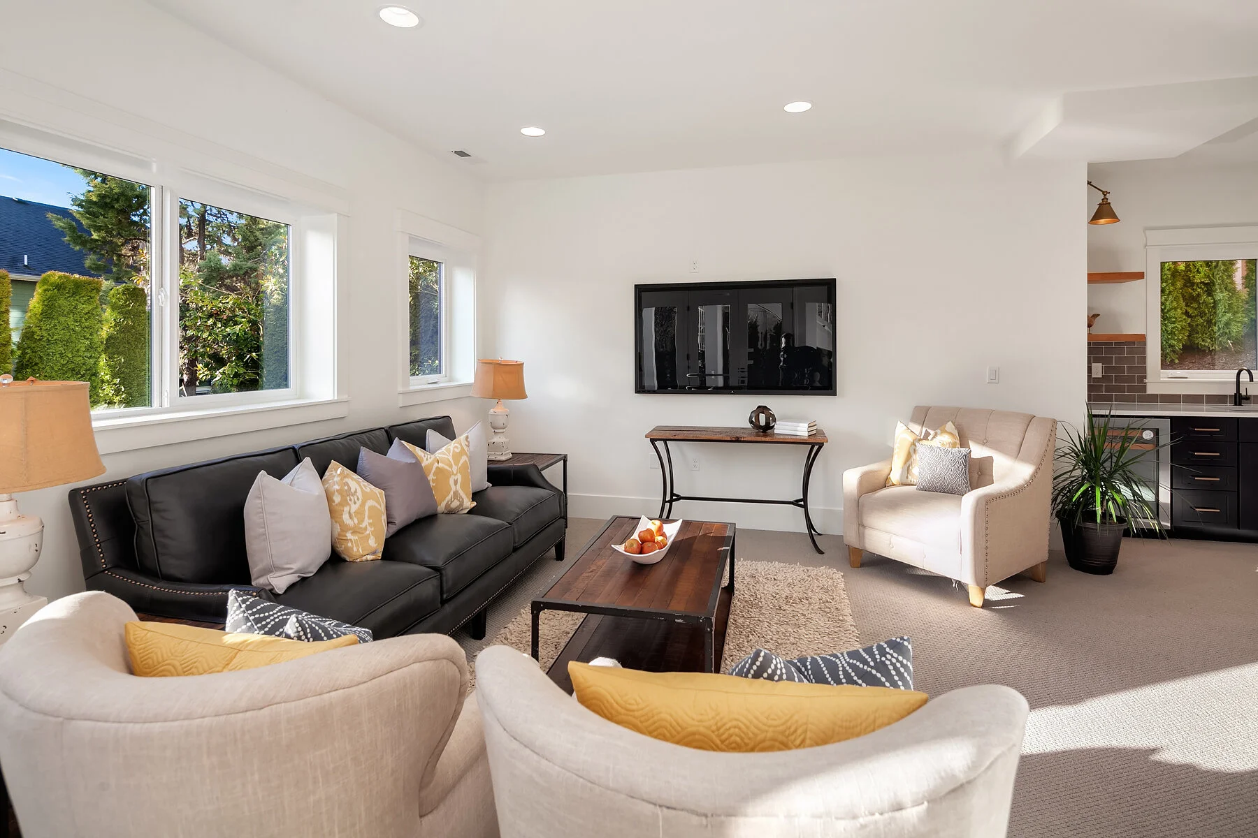 Living room with white walls, large windows, black sofa with yellow and gray pillows, beige armchairs, wooden coffee table with bowl of oranges, wall-mounted TV, side table with lamp, and kitchen area with black cabinets and greenery outside.