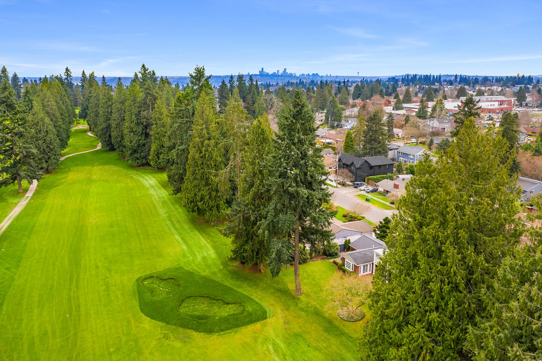 Aerial view of a golf course with lush green fairways, tall trees, and residential houses in the background, with a city skyline visible in the distance under a blue sky.