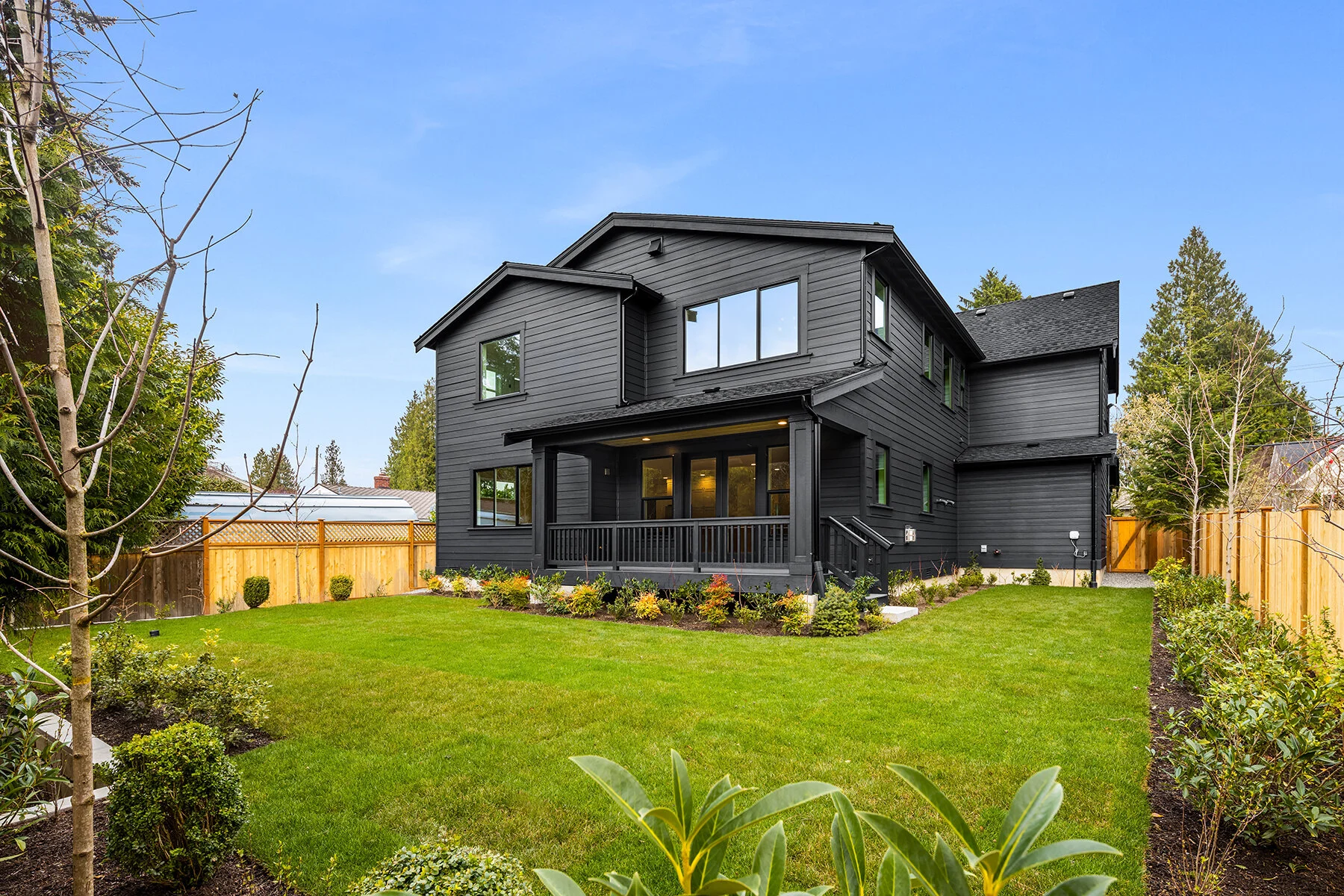 A modern two-story black house with a large backyard, surrounded by green grass and a garden with bushes and trees, enclosed by a wooden fence.