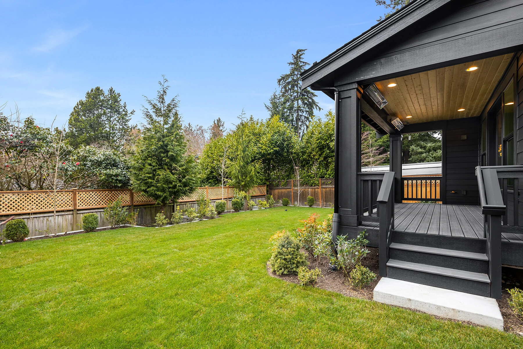 View of a backyard with a green lawn, small bushes, and a wooden privacy fence, next to a black wooden porch with steps leading up to it, under a blue sky.
