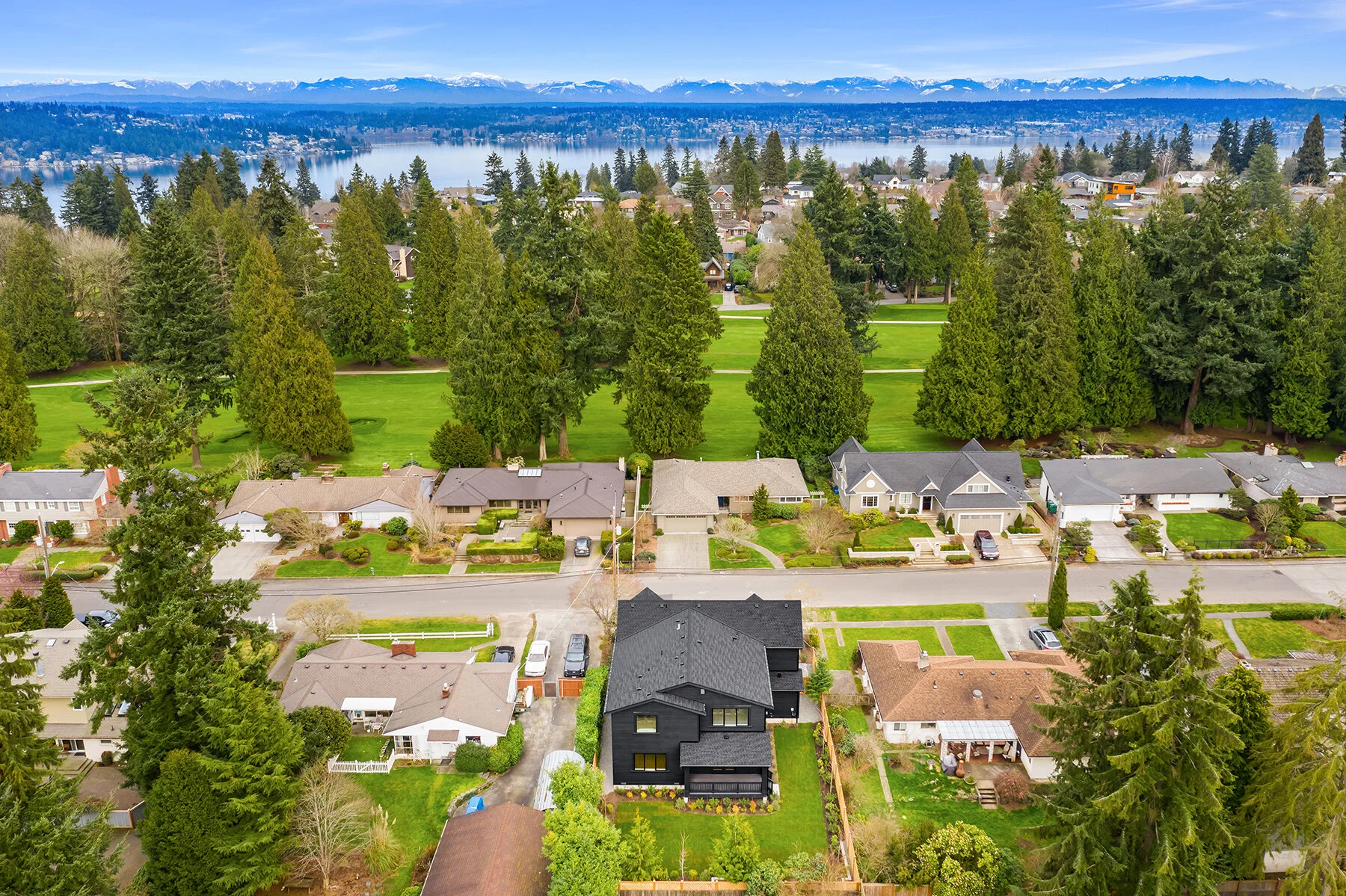 Aerial view of a suburban neighborhood with houses, trees, and a park with a lake and mountains in the background.