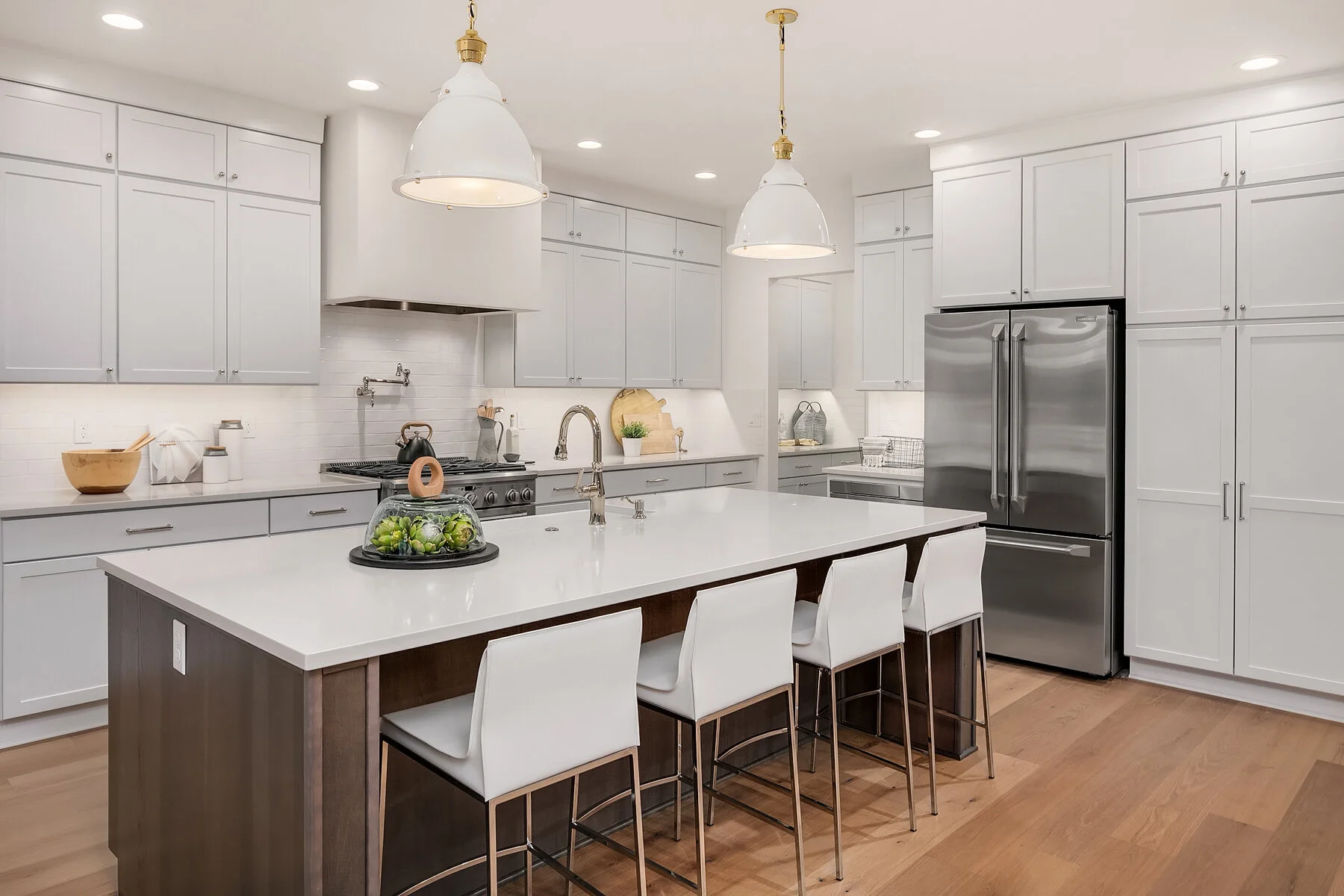 Modern kitchen with white cabinets, a large central island with white countertop, four white chairs, stainless steel refrigerator, and wooden floor, illuminated by ceiling lights and pendant lamps.