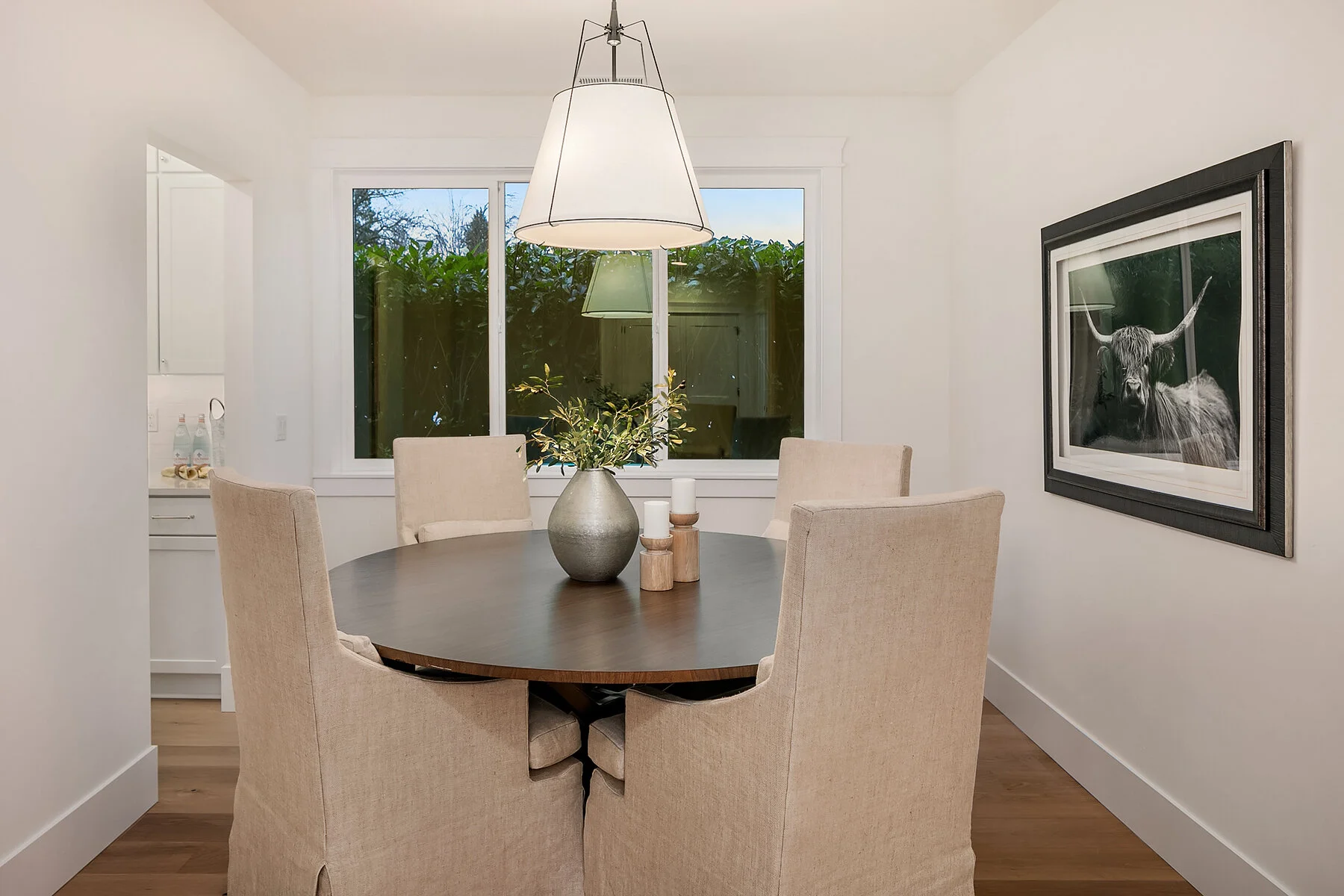 Dining room with a round wooden table surrounded by six beige upholstered chairs, a large gray vase with green foliage on the table, candles on small wooden holders, a large window with greenery outside, a framed painting of a highland cow on the wal