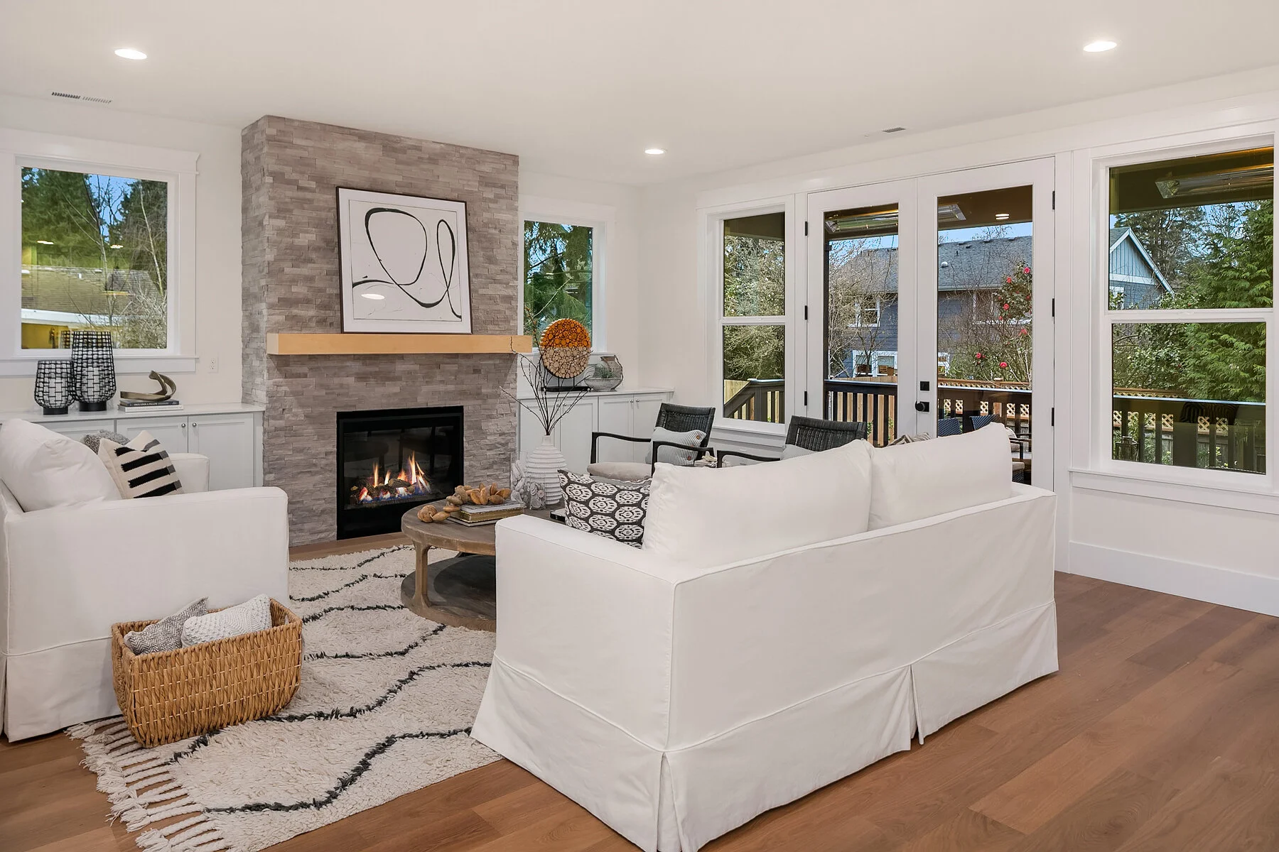 Living room with white sofas, a fireplace with gray stone surround, a wooden coffee table, black and white pillows, large windows and a door leading outside, with plants and houses visible outside.