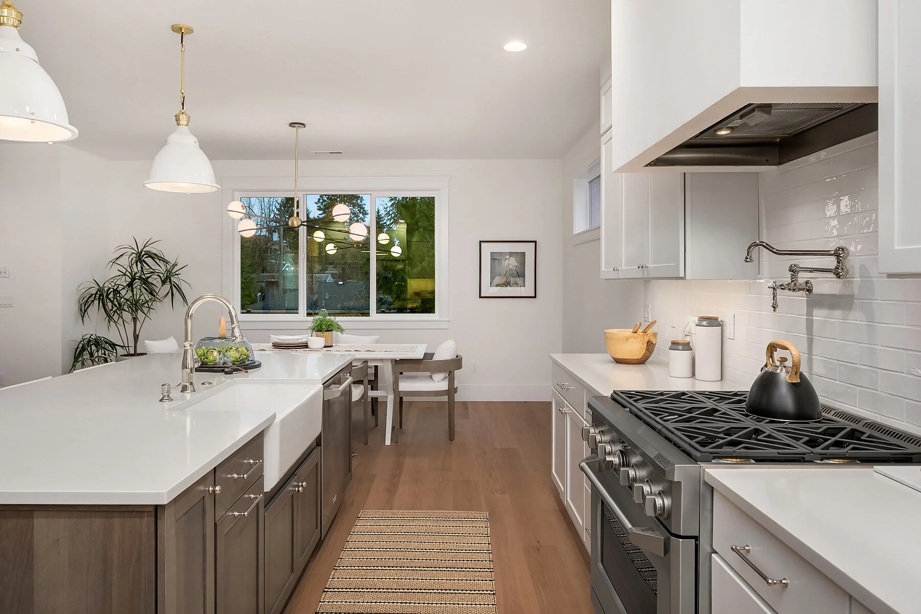 Modern kitchen with white countertops, white cabinets, stainless steel appliances, and wooden flooring. There is a window and a dining area with chairs and a table in the background. Decorative lighting fixtures hang from the ceiling.