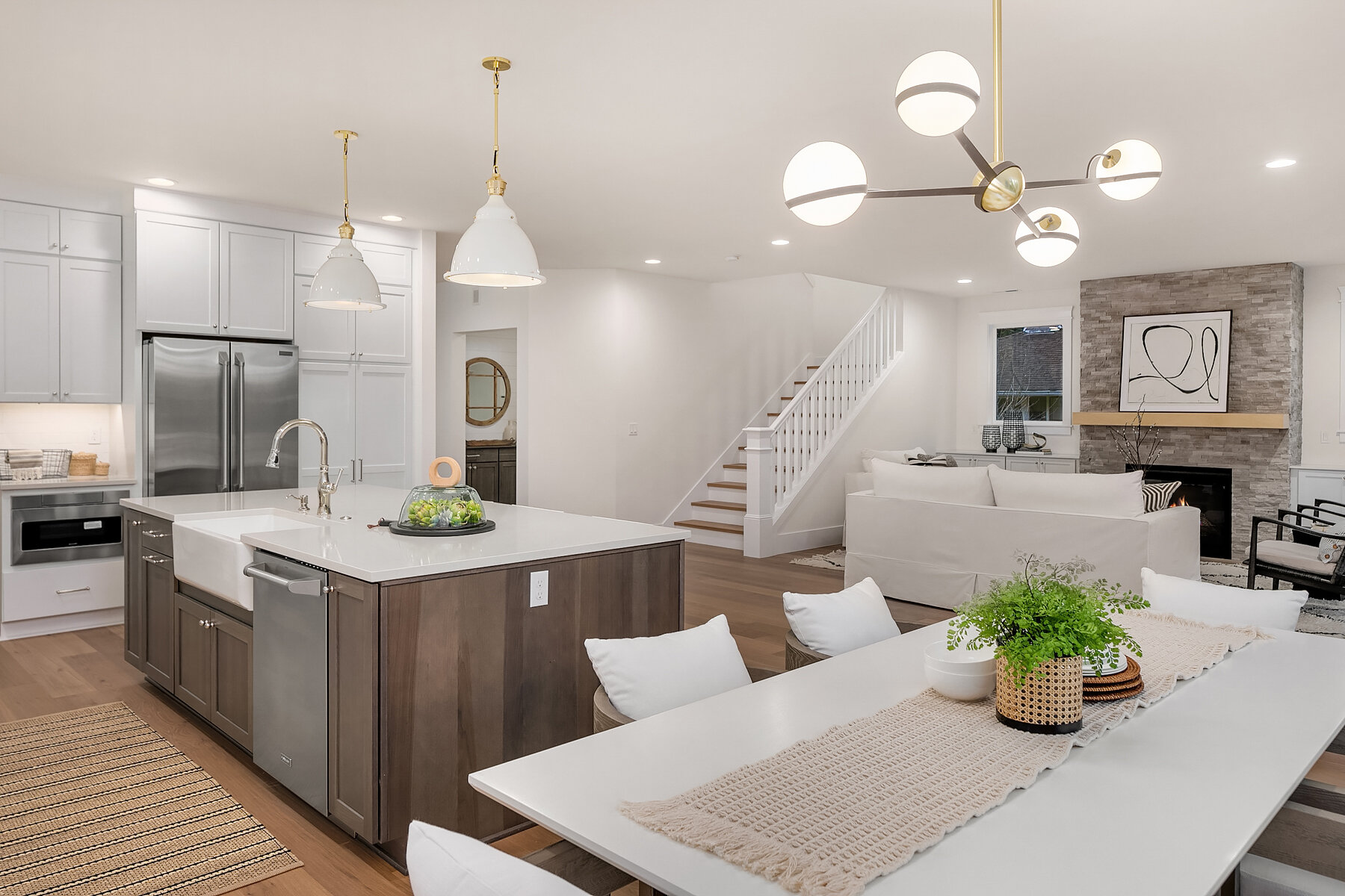 Open-concept kitchen and living room with white and wood accents, modern lighting fixtures, fireplace with gray stone surround, and a dining table decorated with a green plant and neutral cloth.