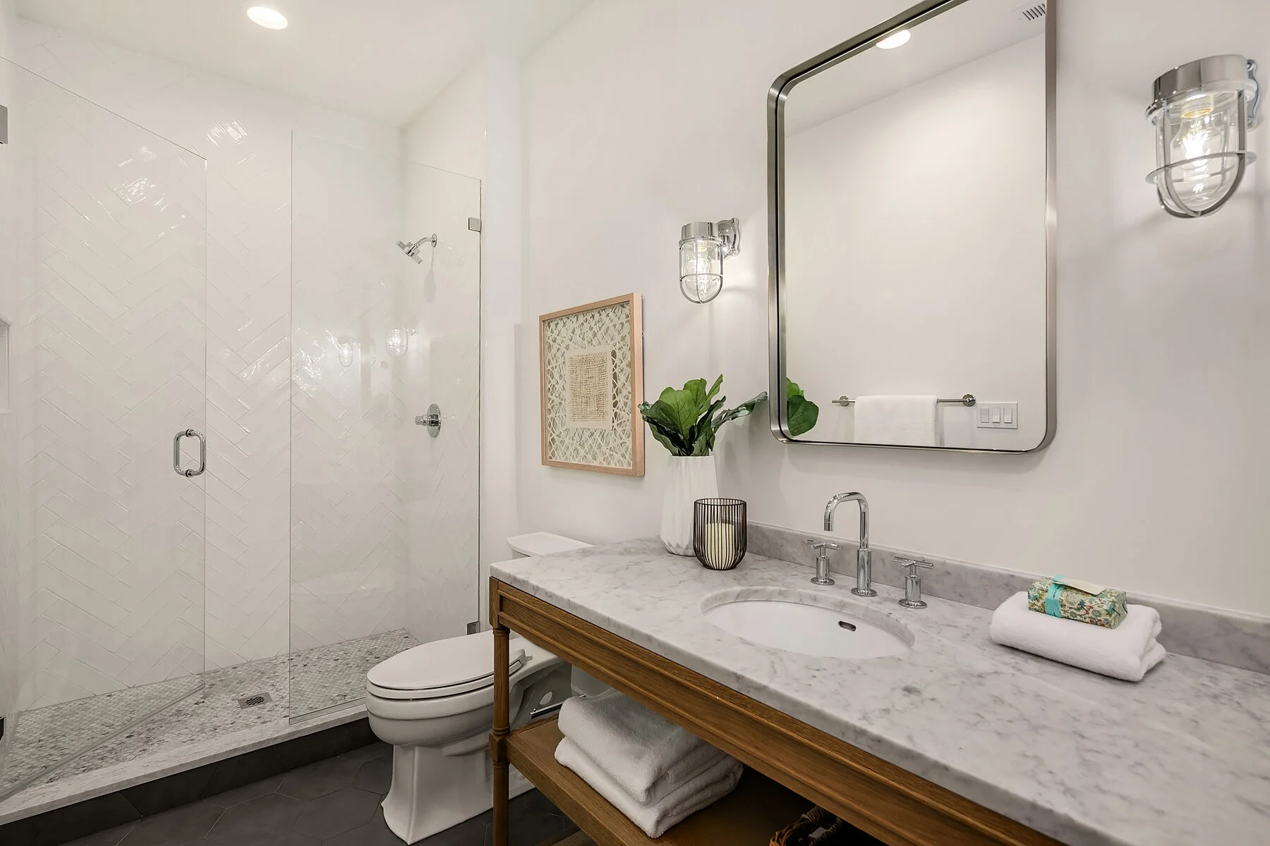 Modern bathroom with walk-in glass shower, marble countertop vanity with framed mirror, potted plant, framed art, towels, and wall-mounted light fixtures.