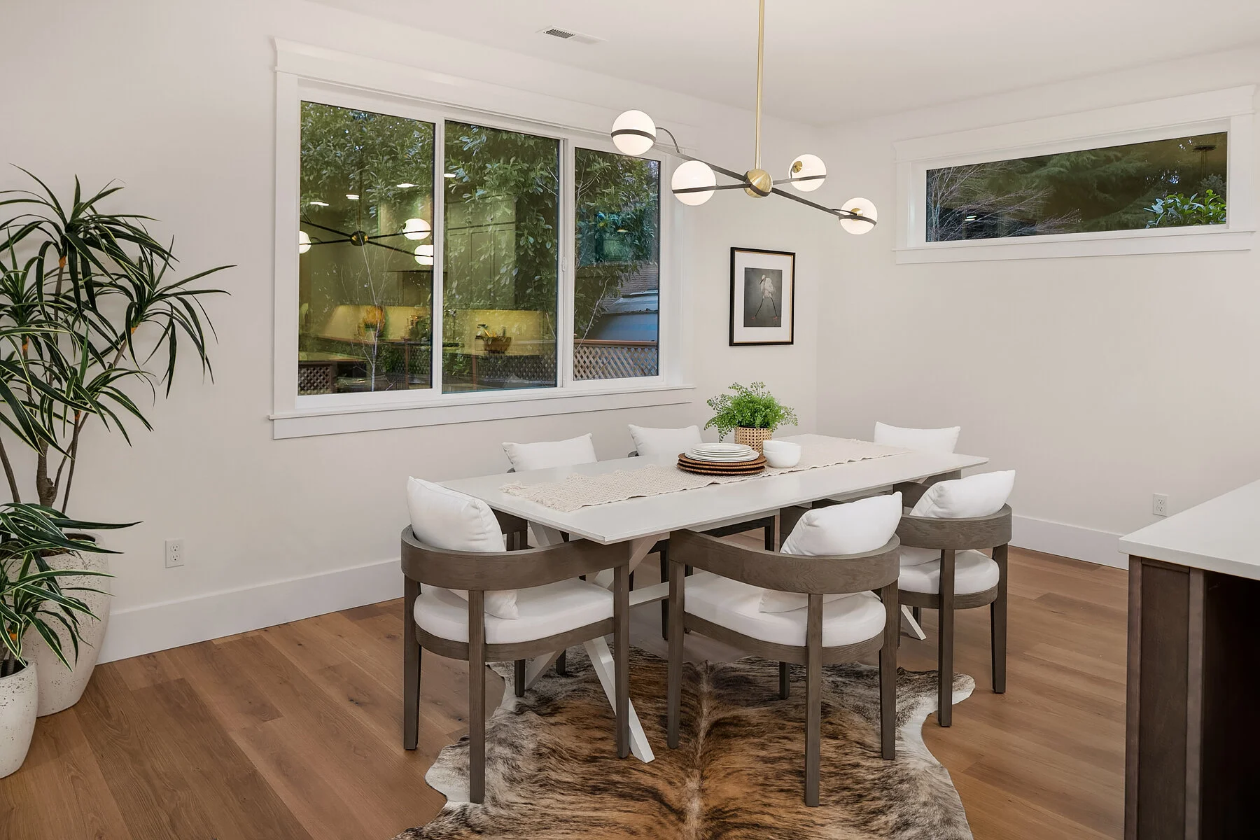 A modern dining room with a white table, six cushioned chairs, a centerpiece plant, and a hanging light fixture. Large window with an outdoor view and woods inside the room, with hardwood floors and a cowhide rug.