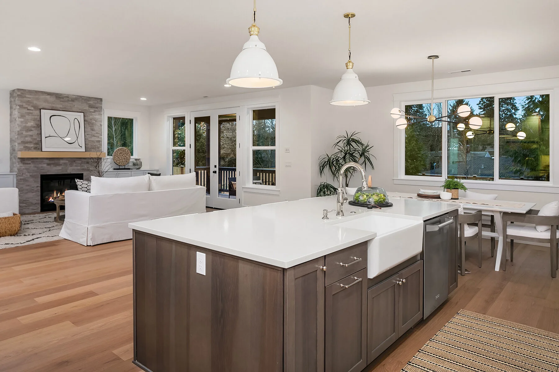 A modern open-concept kitchen and living room with a large kitchen island featuring a farmhouse sink, white countertops, and dark wood cabinets. The living room has a white sofa, a fireplace, and wall art. There are pendant lights above the kitchen i
