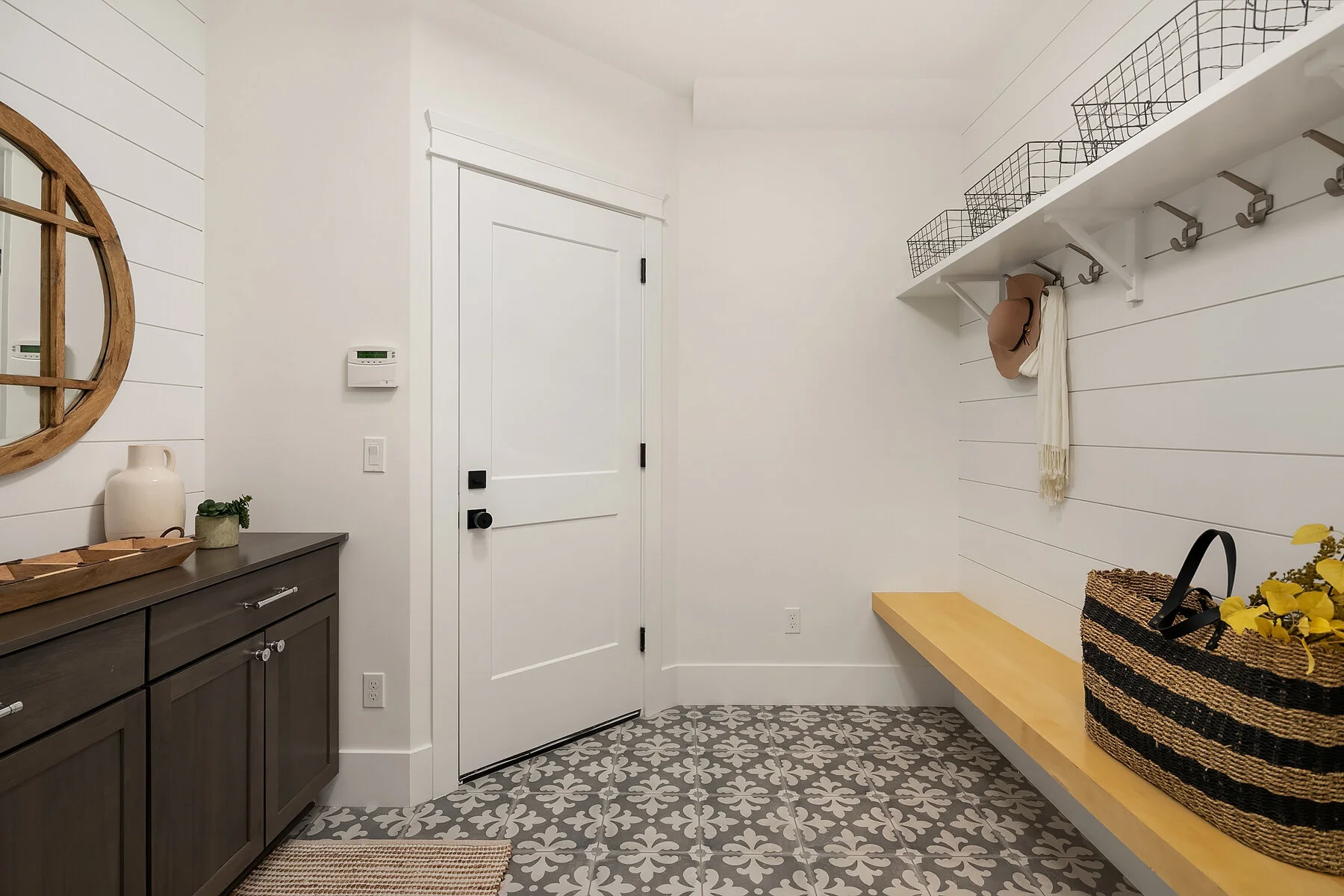 Entryway with white walls, patterned gray and white floor tiles, a dark gray cabinet on the left, a white door in the center, a wooden bench on the right, and wall-mounted storage baskets and hooks.